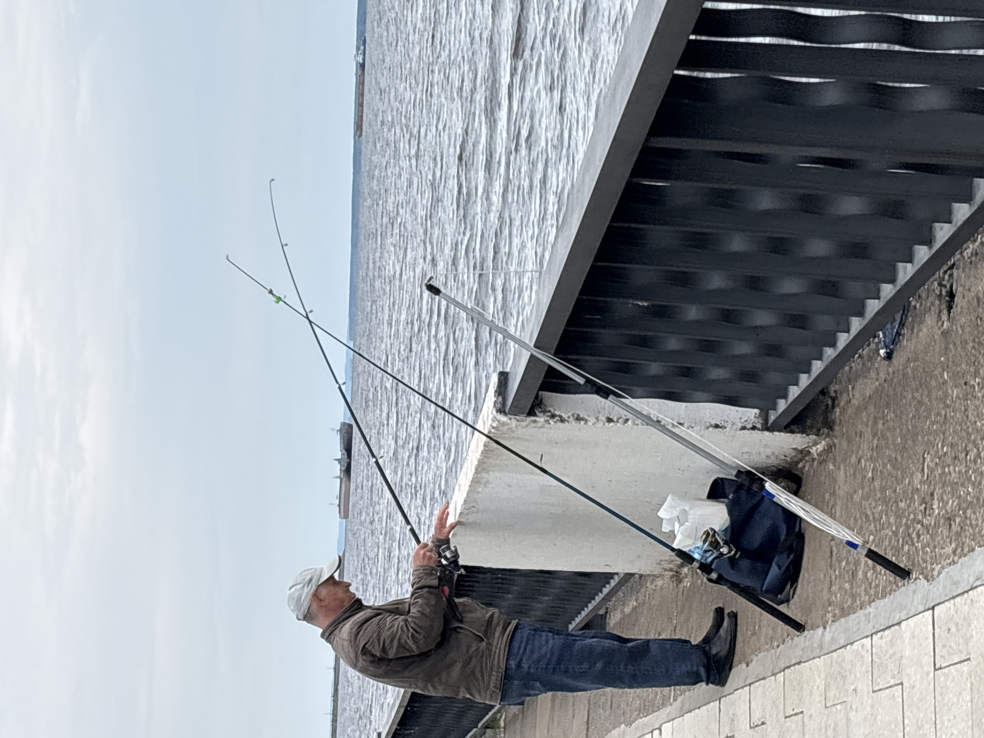 The image shows a man fishing by a large body of water, likely the Kama River in Chaikovsky, Russia. The man, appearing middle-aged, stands in profile on the left side of the frame, facing right towards the water. He wears a light-colored baseball cap, a brown or khaki jacket, blue jeans, and black shoes. He is holding a fishing rod with his left hand near the lower part of the rod and his right hand raised, appearing to be in the process of casting or adjusting his line.
Two additional fishing rods are visible; one is propped up next to a concrete structure near the man, and a longer pole, possibly a net or gaff, leans against the same structure further to the right. A blue bag and some white crumpled items are placed on the ground next to the concrete structure. The water surface, with gentle ripples, extends across the middle and background of the image. Above the water and to the right, a dark metal structure with horizontal bars, possibly part of a bridge or an elevated walkway, is visible. The sky is a clear light blue with sparse clouds, indicating fair daytime weather. No text is visible in the image.