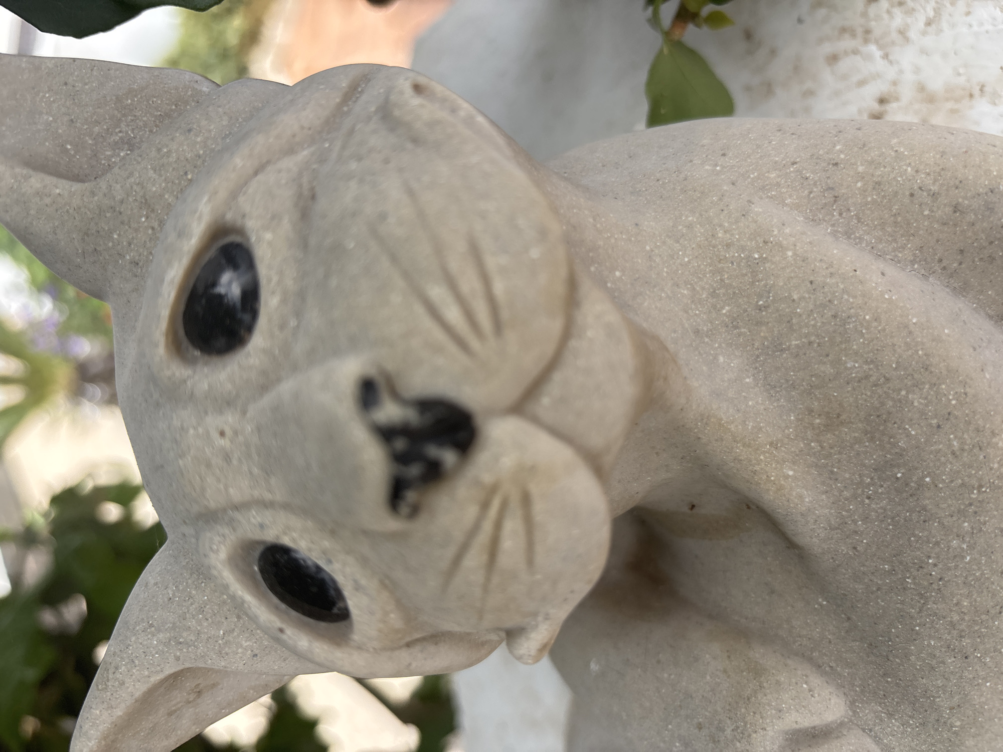 This is a close-up, slightly blurry, tilted photograph of a light-colored, speckled stone sculpture of a rabbit. The sculpture is positioned at an angle, with its head tilted downwards and towards the viewer. The rabbit has large, dark, round eyes, a small black nose, and faint incised lines on its muzzle representing whiskers. Its ears are rounded and sit prominently on top of its head. The texture of the stone is visible, showing a grainy, speckled surface. The background is out of focus and appears to be an outdoor setting with green foliage and some lighter, possibly white, elements. There is no visible text in the image. The lighting suggests it is daytime, but the weather is not discernible. The overall impression is of a decorative garden statue.