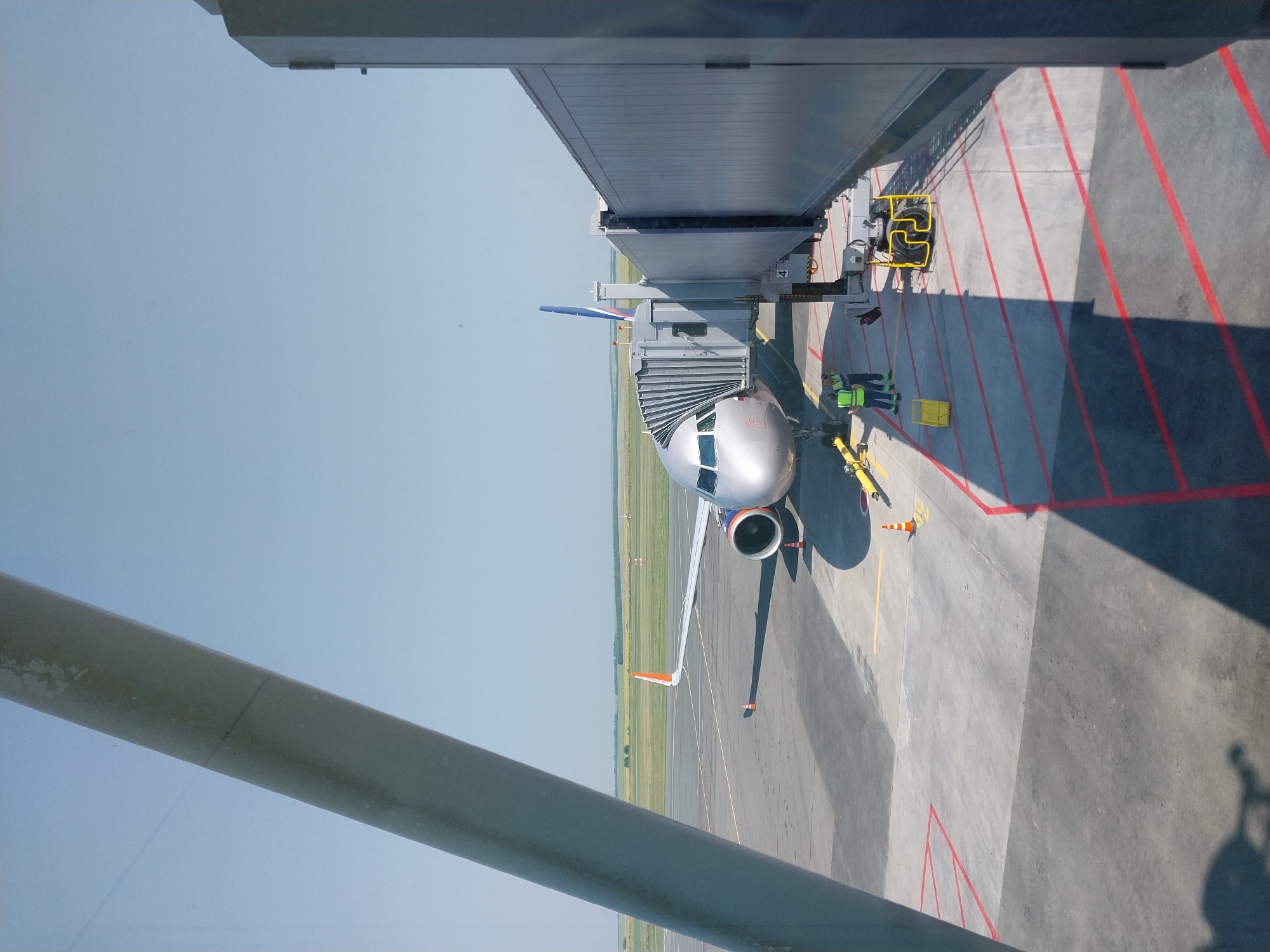 The image shows an airplane parked at an airport gate, connected to a jet bridge. Two airport workers in high-visibility vests are standing near the jet bridge. The scene is set on a sunny day at an airport, with a clear blue sky overhead and a vast green field visible in the distance. The airplane is a silver passenger jet with orange wingtips. The jet bridge is gray and appears to be extended to meet the aircraft. The tarmac is marked with red lines. The image appears to have been taken from inside an airport terminal or another aircraft. The location is identified as Orenburg, Russia.