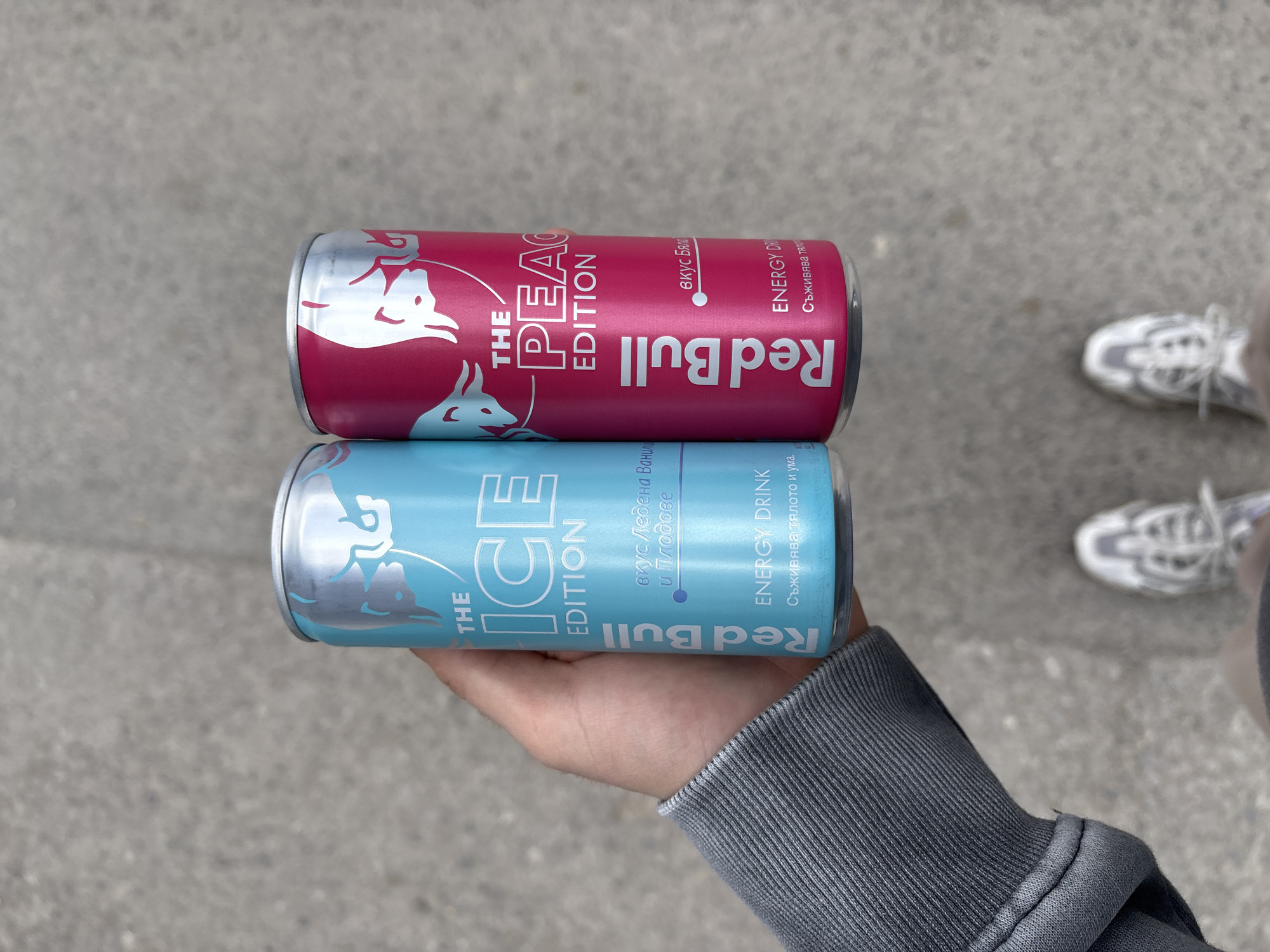 The image shows two cans of Red Bull energy drink being held in a person's hand. The cans are stacked on top of each other. The top can is a "Peach Edition" Red Bull, and the bottom can is an "Ice Edition" Red Bull. The cans have Bulgarian text on them. The person is wearing a gray hooded sweatshirt. The background is a gray concrete surface. Parts of white sneakers can be seen. The picture was likely taken outside. There are no specific details to determine if this image was taken in the city of Sofia, Bulgaria.