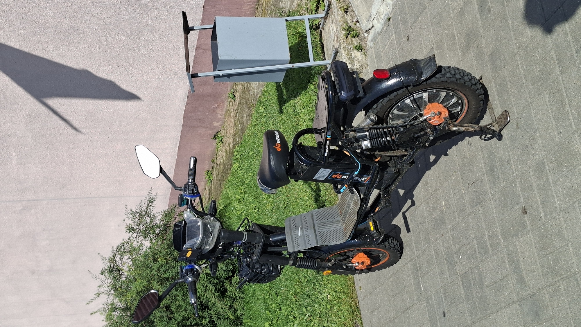 The image depicts an electric scooter parked on a paved walkway in front of a pinkish-brown building. A green bush is to the left of the scooter, and a gray trash bin stands to the right. The scooter is black with orange accents on the wheels. It has a basket in the front, a gray footrest, a black seat with an orange logo, and a visible battery pack. The scooter is standing on its kickstand. The overall scene is outdoors, in daylight, under a clear sky, as indicated by the shadow of a large object on the building's wall. The location is identified as Myadzyel, Belarus. There are no people visible in the image, and no discernible actions are taking place.