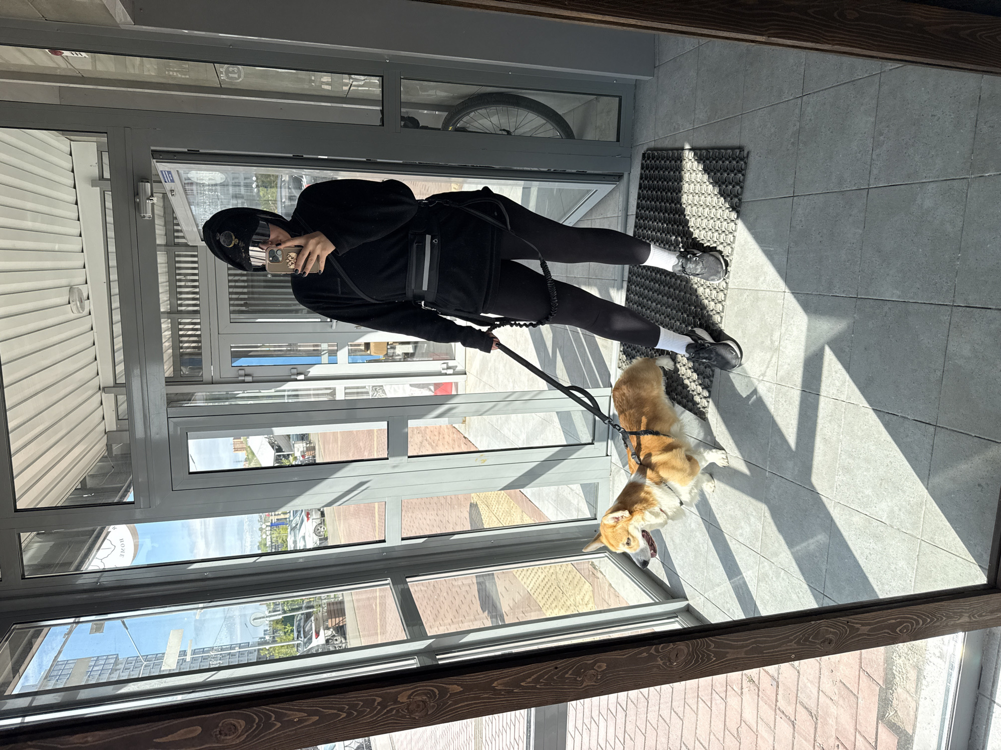A person dressed in black, including a hoodie and leggings, is standing in a doorway taking a selfie with a phone. They are wearing white socks and dark sneakers. The person is holding a black leash attached to a corgi dog that is standing near the door. The doorway is composed of glass panes and metal frames. Outside the glass, there are reflections of buildings and the sky, suggesting an urban setting. A doormat is visible at the person's feet. Based on the reflection in the glass, the location is Krasnoyarsk, Russia. A partial view of a bicycle is visible through a window above the person.