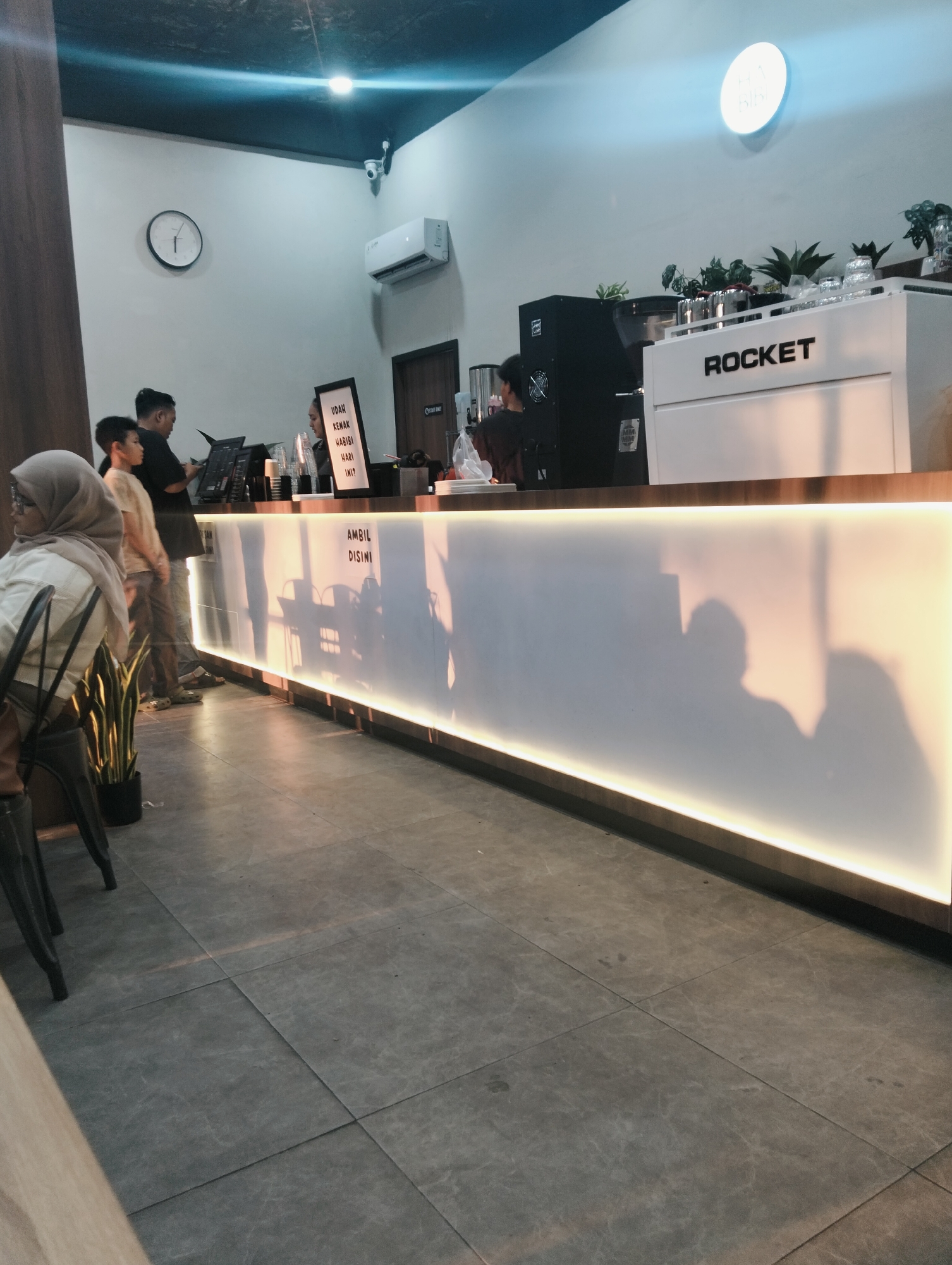 The image depicts the interior of a cafe in the city of Medan, Indonesia. A long, white counter with a wooden top runs across the mid-ground, illuminated from below by a warm light. The counter has a translucent front that casts shadows of people onto it. On the counter, various items are visible, including what appears to be a coffee machine labeled "ROCKET" and some potted plants. Behind the counter, a cashier, likely a woman, is interacting with a customer, a man, who is looking at a laptop. Another man and a younger boy are standing nearby, seemingly waiting. In the foreground, a woman wearing a hijab is seated at a table to the left, partially visible. The floor is tiled with large, gray square tiles. A clock and an air conditioner unit are mounted on the light-colored wall in the background. A security camera is also visible on the ceiling. The overall atmosphere suggests a modern and casual cafe setting.
