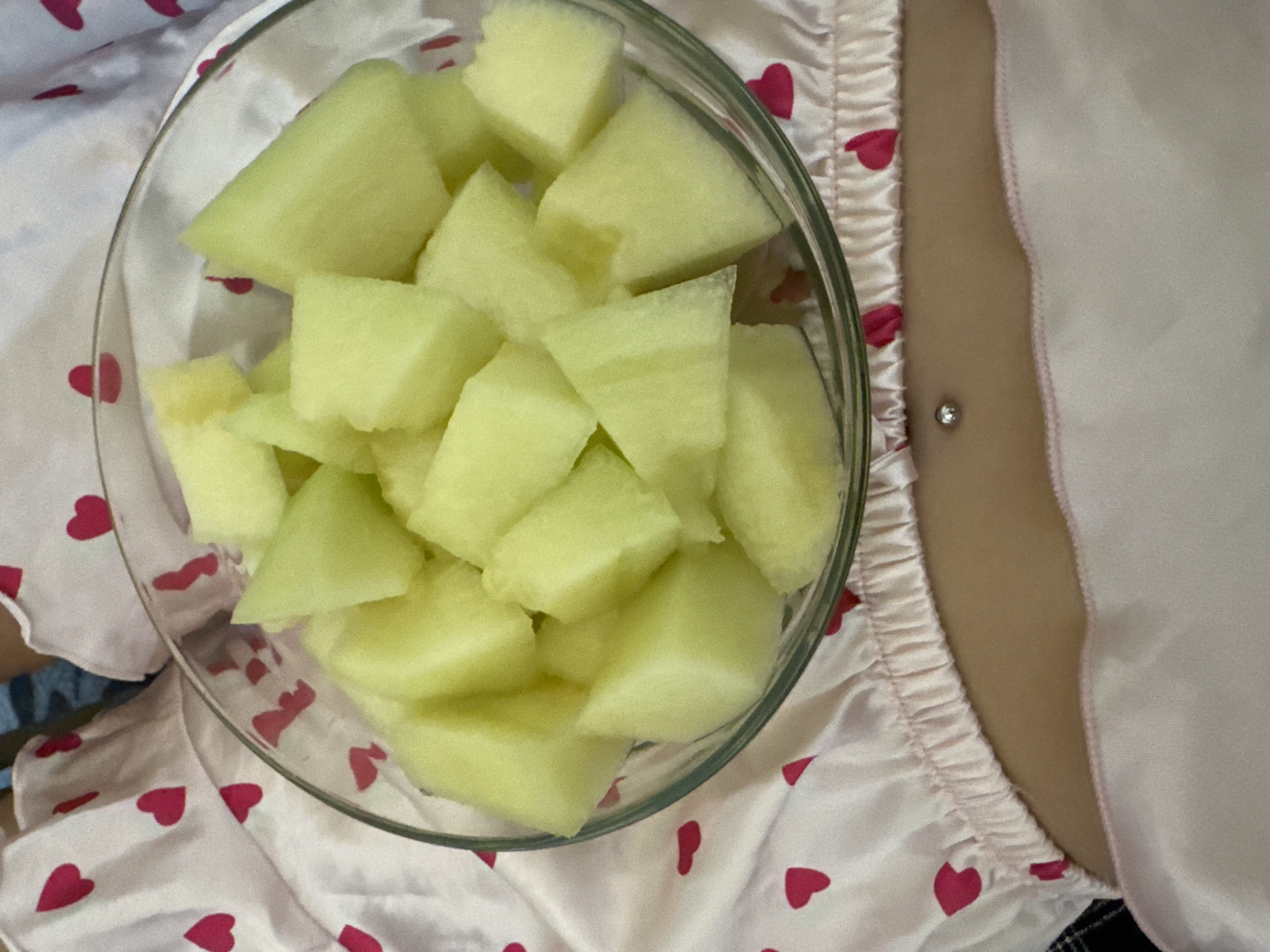 An indoor scene, set in Plovdiv, Bulgaria, displays a close-up view of a person's torso. A clear glass bowl, positioned towards the top of the frame, is filled with numerous uniformly diced pieces of pale green fruit, consistent with melon. Below the bowl, a light-colored fabric, possibly part of pajamas or loungewear, is visible, patterned with scattered red hearts and featuring an elasticized, gathered hemline. Directly beneath this fabric, a section of bare skin is exposed, revealing a navel piercing adorned with a small, spherical metallic bead. Further down, a smooth, light-pink fabric, likely another garment or bedding, is visible, partially obscured by the foreground. The overall lighting is soft and even.