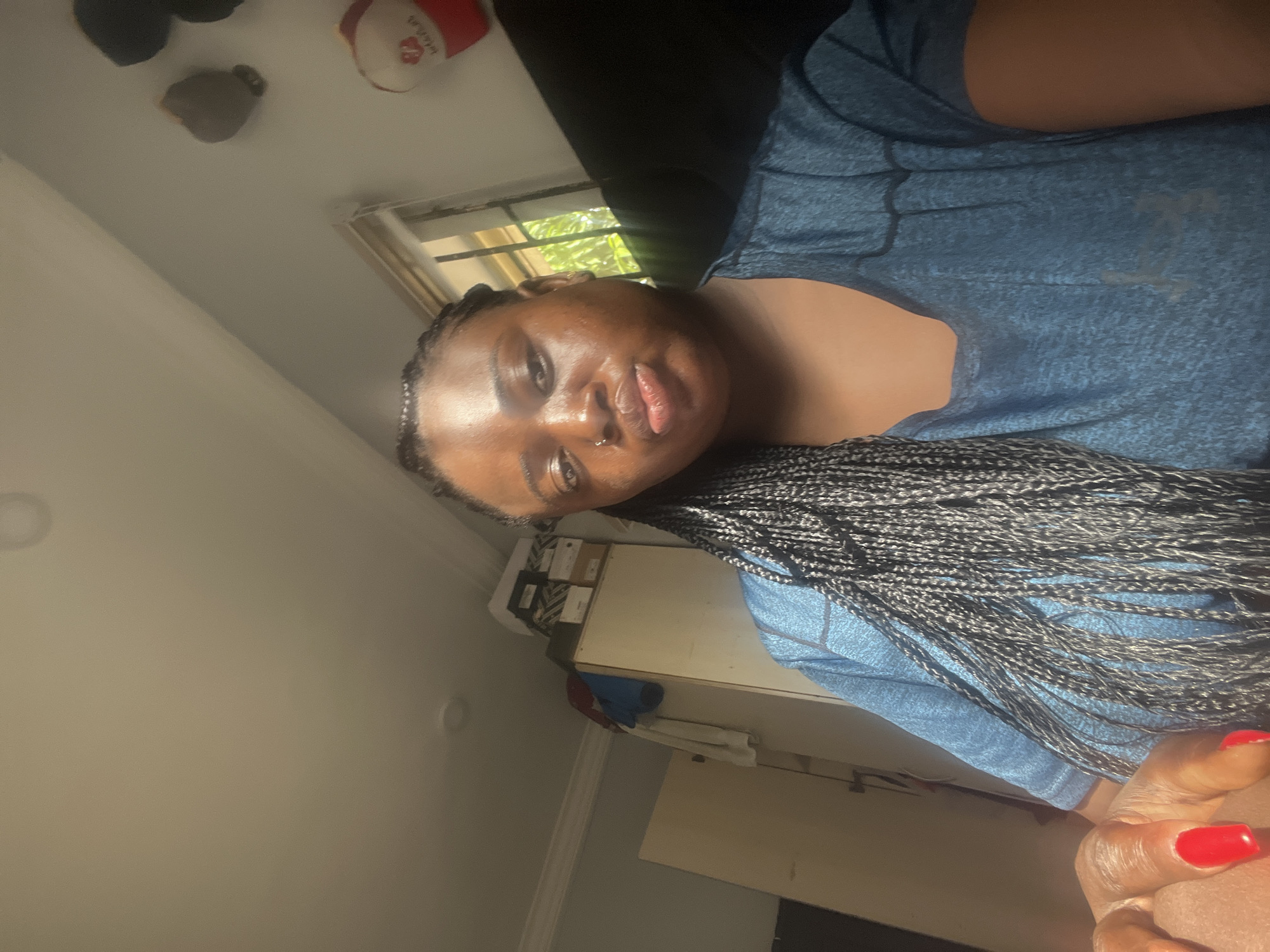 A person is lying on a dark surface. They are wearing a blue shirt and have braids. One of their hands is visible with red nail polish on the fingers. The location appears to be indoors, with a window visible and some objects on a shelf in the background. The city of Dushepe, Nigeria is not evident in the photo.