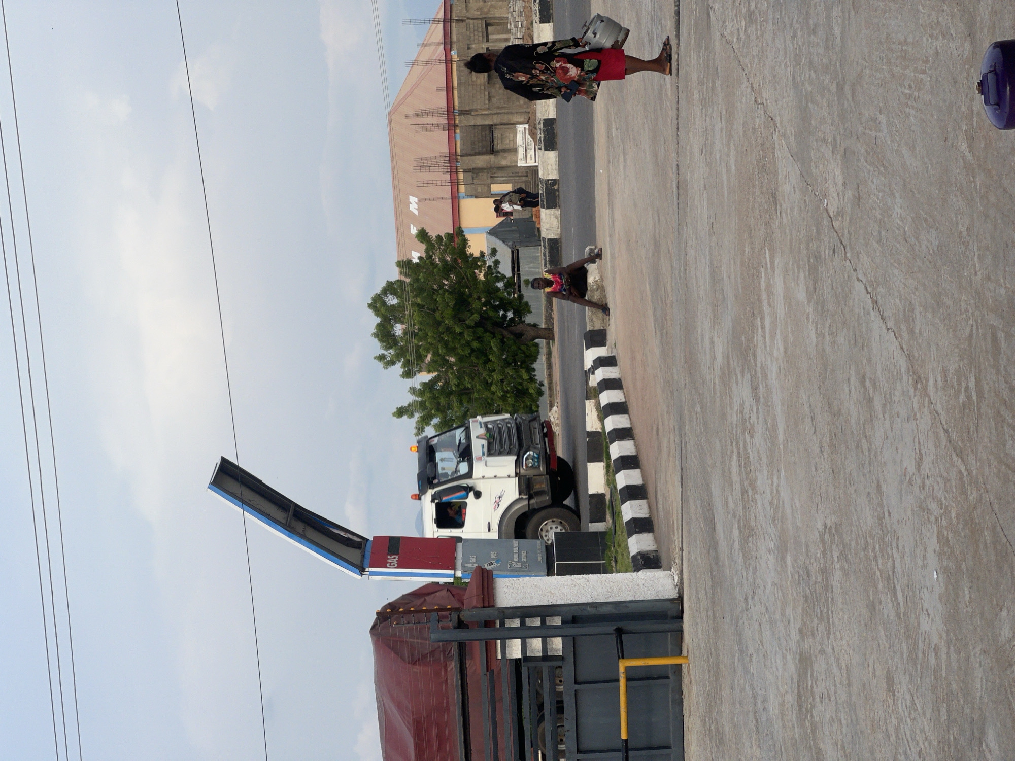 An exterior medium shot captures a gas station with a white truck dispensing fuel under a cloudy sky in Jalingo, Nigeria. A large, red-covered truck is parked to the left, with a gray gate and yellow barrier in the foreground. A gas pump structure with "GAS" displayed prominently stands between the trucks. In the middle ground, a young man sits on a concrete curb, facing away from the camera. To the right, a woman walks away from the camera, carrying a metal container in her right hand. Behind them, a building under construction with exposed rebar and a red-tiled roof is visible. The ground is paved concrete, with black and white striped markings on the curb. Power lines are strung across the sky.