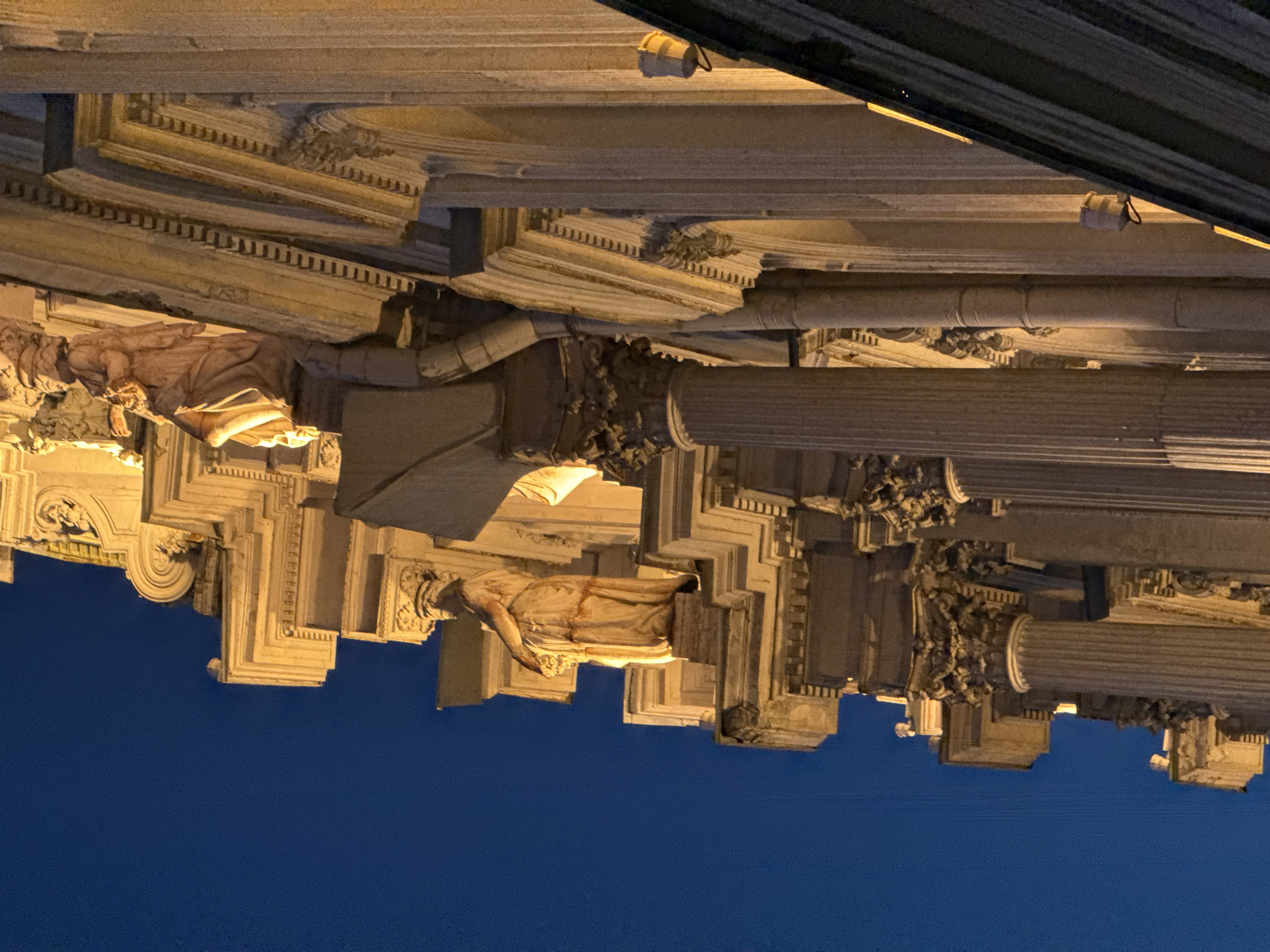 The image shows an architectural detail of the Singer House, a historic building located in Saint Petersburg, Russia. The perspective is from below, looking up at the facade at dusk. The building features ornate classical architecture with columns, statues, and decorative carvings, illuminated by artificial light. The sky behind is a deep blue, indicating twilight. One statue of a woman in flowing robes is clearly visible on a ledge, holding what appears to be a bouquet or cornucopia. Another, more partially obscured statue is visible further up and to the right. The building's facade is made of stone, with a warm, golden hue from the lighting.