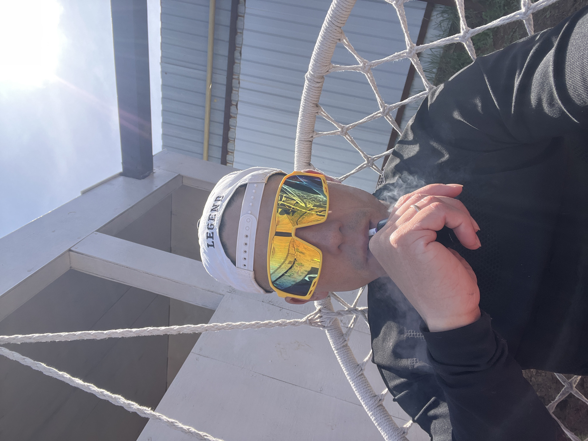 A person in sunglasses and a "Legend" hat is sitting in a rope chair and smoking. The background includes a building with a metal roof and white support structures. The location is indeterminate; there is no information to determine it is in Chaikovsky, Russia.