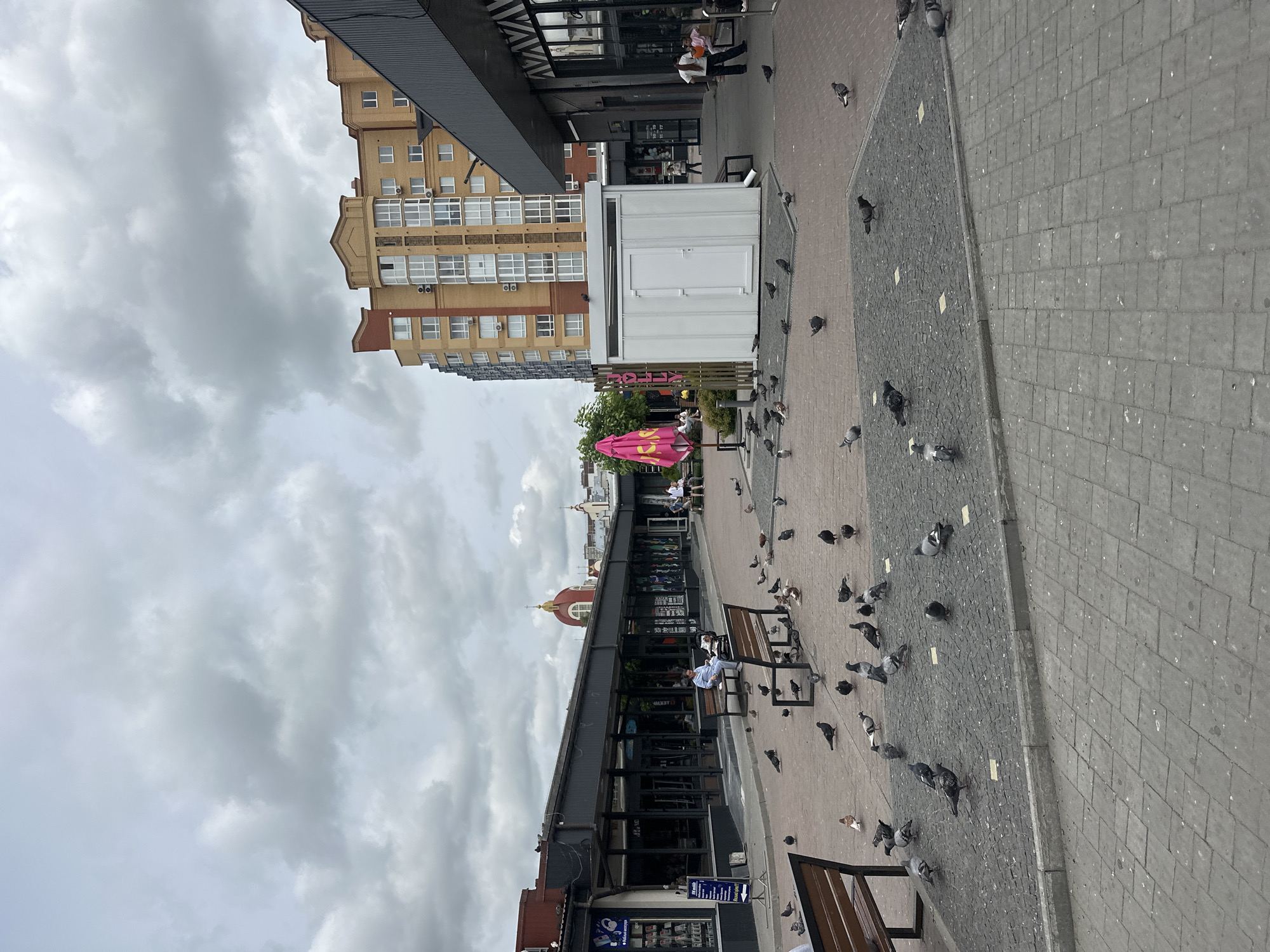 The image shows a public square in Ternopil, Ukraine, with a paved ground area filled with pigeons. Several benches are placed around the square, with some people sitting on them. To the left of the square are a row of shops with glass storefronts. In the background, there is a modern apartment building and a building with a red dome, possibly a church. The sky is cloudy.