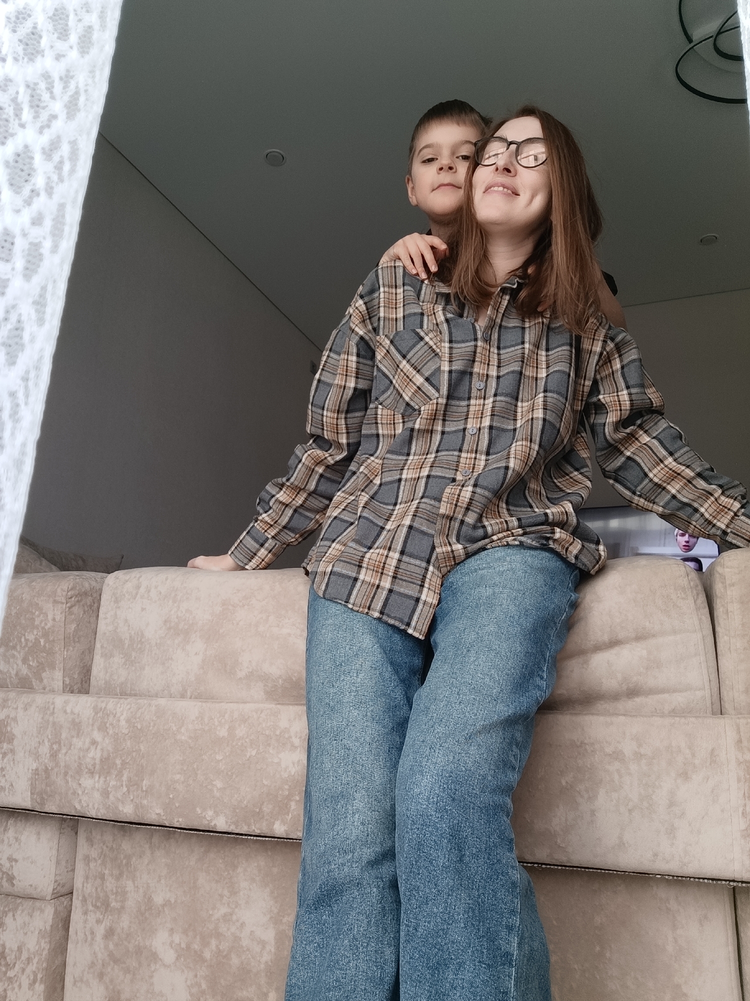 A woman with brown hair and glasses, wearing a plaid shirt and jeans, is sitting on a beige couch. A young boy stands behind her, with his hand on her shoulder. The location is in Chaikovsky, Russia, inside a home. A white lace curtain is visible to the left. In the background, there is a gray wall and ceiling, and light fixtures. A television screen can be seen behind the woman.