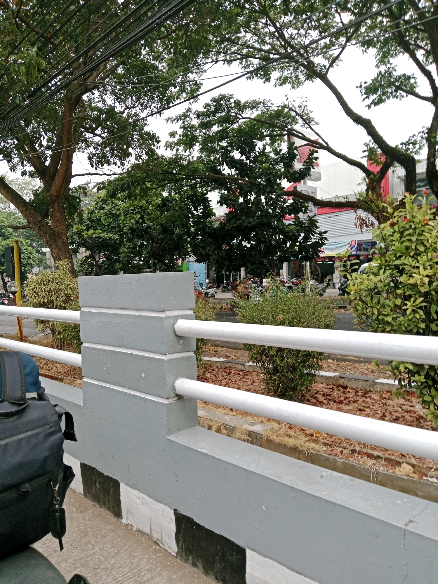 A low-angle, eye-level outdoor shot shows a grey concrete barrier with white horizontal rails in the foreground, separating a sidewalk from a vegetated area. A black backpack is visible on the left side, resting on what appears to be a motorcycle seat. Beyond the barrier, trees with dense green foliage dominate the midground, with power lines strung above them. In the background, buildings with signs and parked motorcycles line a street. The scene suggests an urban environment in Muka, Indonesia, with a clear division between pedestrian and vehicular areas, and a focus on infrastructure and greenery.
