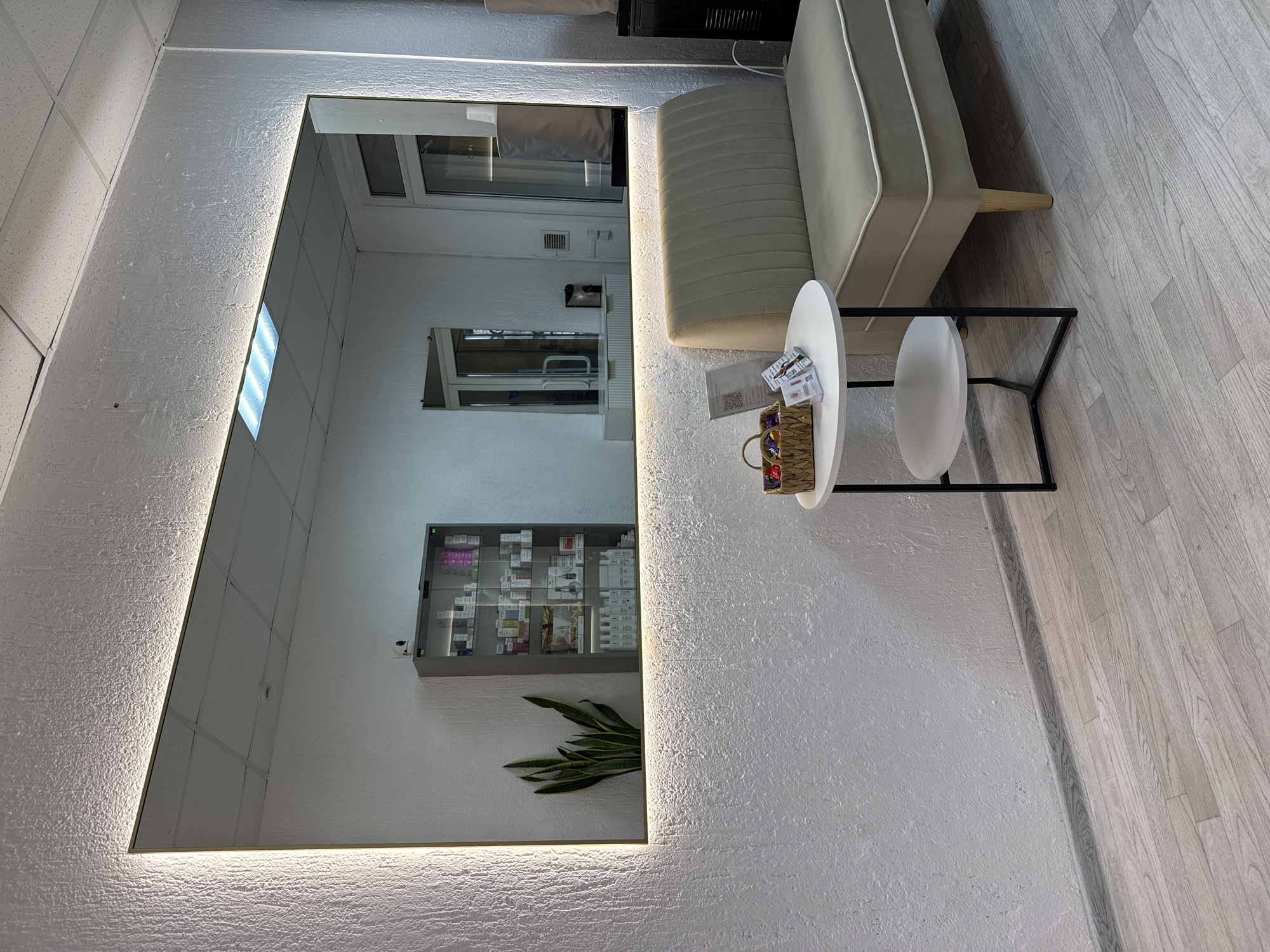 This media file captures an interior scene, likely a waiting area in a commercial establishment such as a salon, clinic, or aesthetic center in Chaikovsky, Russia. No people are visible, and no specific activity is taking place, presenting a static view of a neat and modern space.
The dominant feature on the left is a large, vertically oriented rectangular mirror framed by a warm, glowing light strip. The light illuminates the pronounced, rough texture of the white wall it's mounted on. Reflections in the mirror reveal parts of the room opposite, including a window, a ceiling with white acoustic tiles and linear light fixtures, and a display cabinet filled with numerous small, packaged products, possibly cosmetics or healthcare items. A smaller, darker rectangular mirror is also visible in the reflection. Below the display cabinet, a green potted plant with tall, pointed leaves (likely a snake plant) is visible.
To the right of the mirror, a light grey or beige sofa with a simple, modern design and wooden legs is positioned. In front of the sofa is a small, round white coffee table with a minimalist black metal frame. On the table, there's a small wicker basket holding indistinct colorful items, alongside several pamphlets or cards, one of which displays a visible but unreadable QR code and some unreadable text.
The flooring throughout the visible area is a light-colored laminate or wood-look tile. The overall ambiance is clean, contemporary, and functional, with a neutral color palette. The artificial lighting from the mirror and ceiling fixtures suggests the time of day is not a significant factor, or it's an indoor setting designed to be well-lit regardless of natural light.