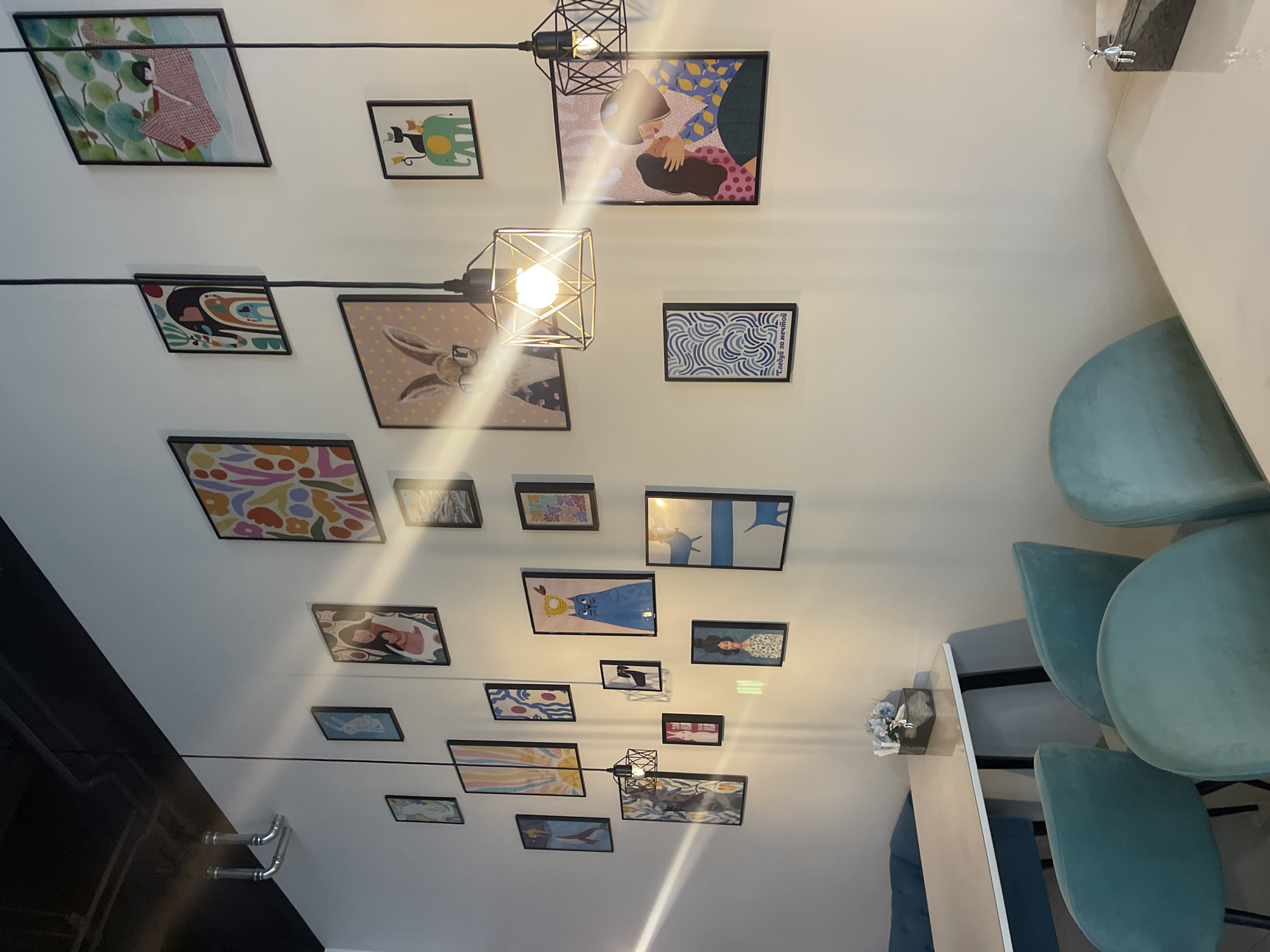The image depicts an interior scene with a wall covered in framed artwork. The artwork includes a variety of colorful illustrations, abstract patterns, and depictions of animals and people. Several pendant lights hang from the ceiling, casting light on the wall and artwork. In the foreground, there are teal-colored chairs and a white table. The table has a rectangular holder with blue flowers and what appears to be a tissue box. The environment suggests a cafe or a dining area. The wall is painted a light color, and a black ceiling is visible at the top of the image. There are no people visible in the image. The artwork is arranged in a gallery-style display. The overall impression is one of a decorated and contemporary space.