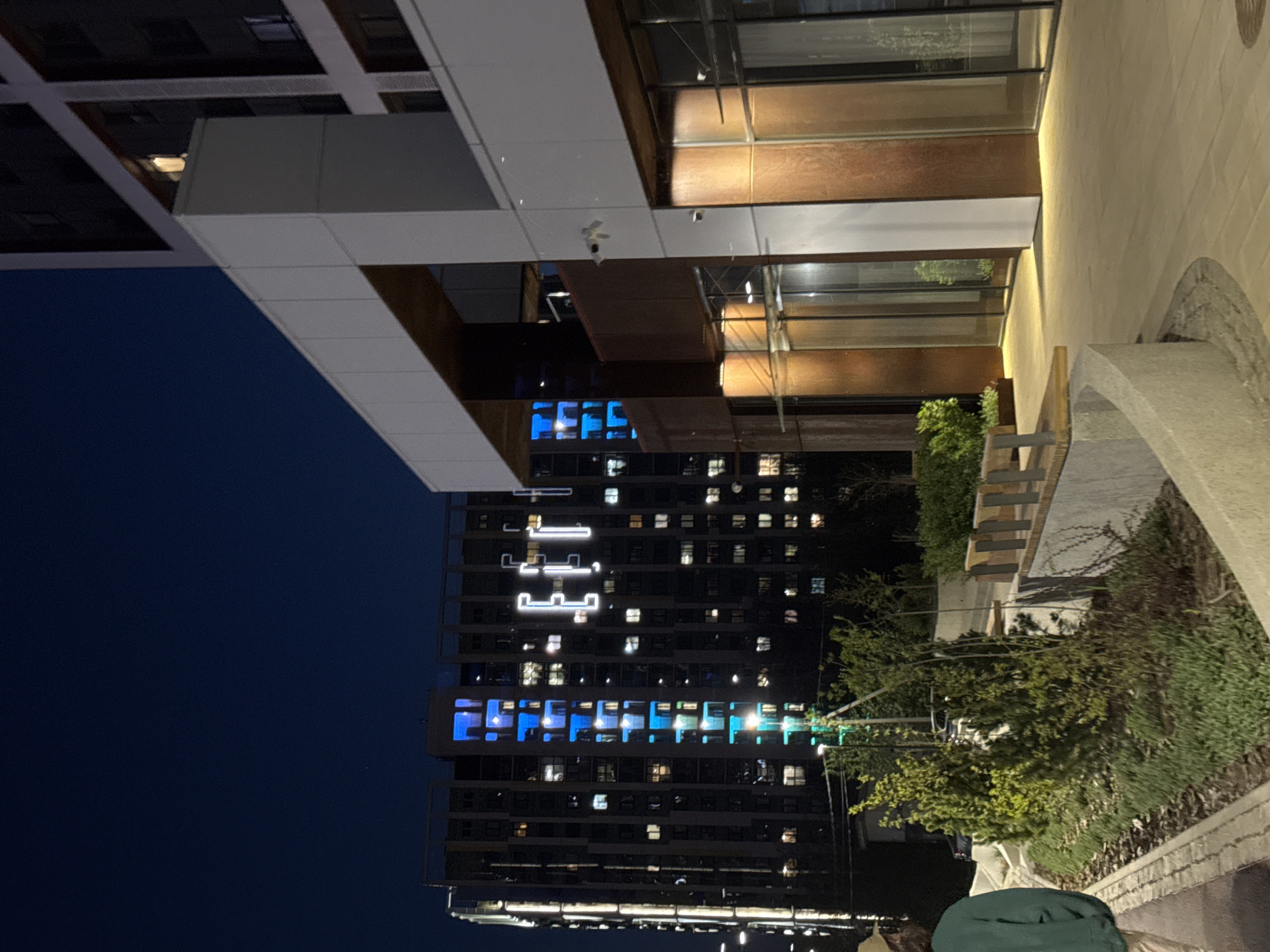 The image depicts an urban scene at night, featuring two prominent buildings. The first is a modern structure with a facade of illuminated glass and concrete. The second is a taller high-rise apartment building in the background adorned with a series of lighted patterns, with a blue and white theme. The location is Izhevsk, Russia, as indicated by the digital window display of the high-rise building. The immediate foreground features a sidewalk with some landscaping and a person. The sky is dark, confirming the nighttime setting.