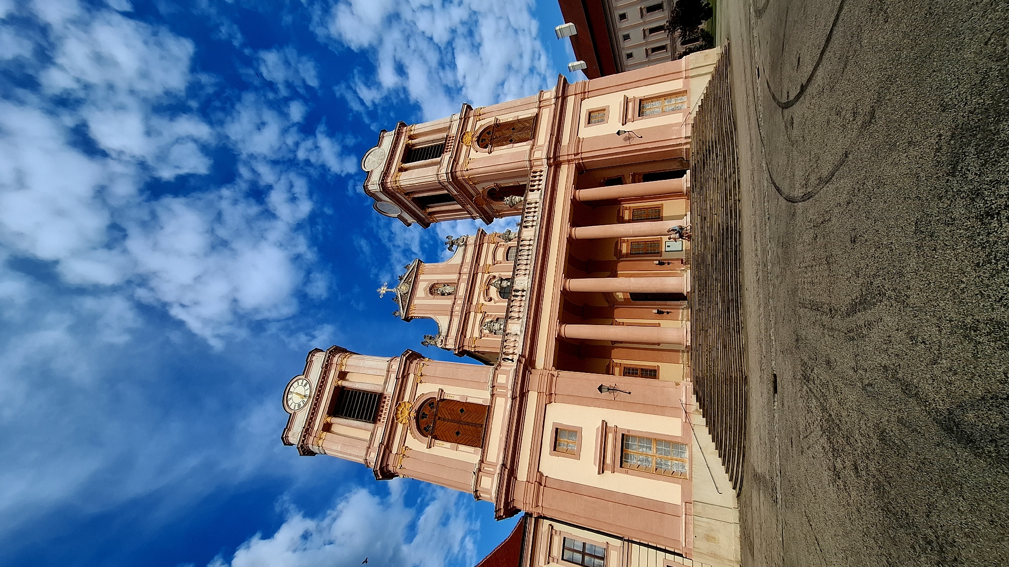 The image shows the Melk Abbey in Furth bei Göttweig, Austria, a magnificent Benedictine monastery. The abbey is a grand Baroque structure with two prominent bell towers, a central facade adorned with statues and a cross, and a portico with large columns. The building is painted in a pale pink and beige color scheme, with contrasting wooden doors and windows. A wide set of stairs leads up to the entrance.
In front of the abbey is a large, paved courtyard. The sky above is a vibrant blue, dotted with fluffy white clouds. The sun is shining, casting shadows and illuminating the architecture.
On the steps of the abbey, two individuals are seated, appearing to be resting or observing the surroundings. The ground in the courtyard shows some tire marks, suggesting recent activity, possibly from vehicles. A lamppost is visible on the left side of the building's base. The overall impression is one of historical grandeur and a peaceful, sunny day.