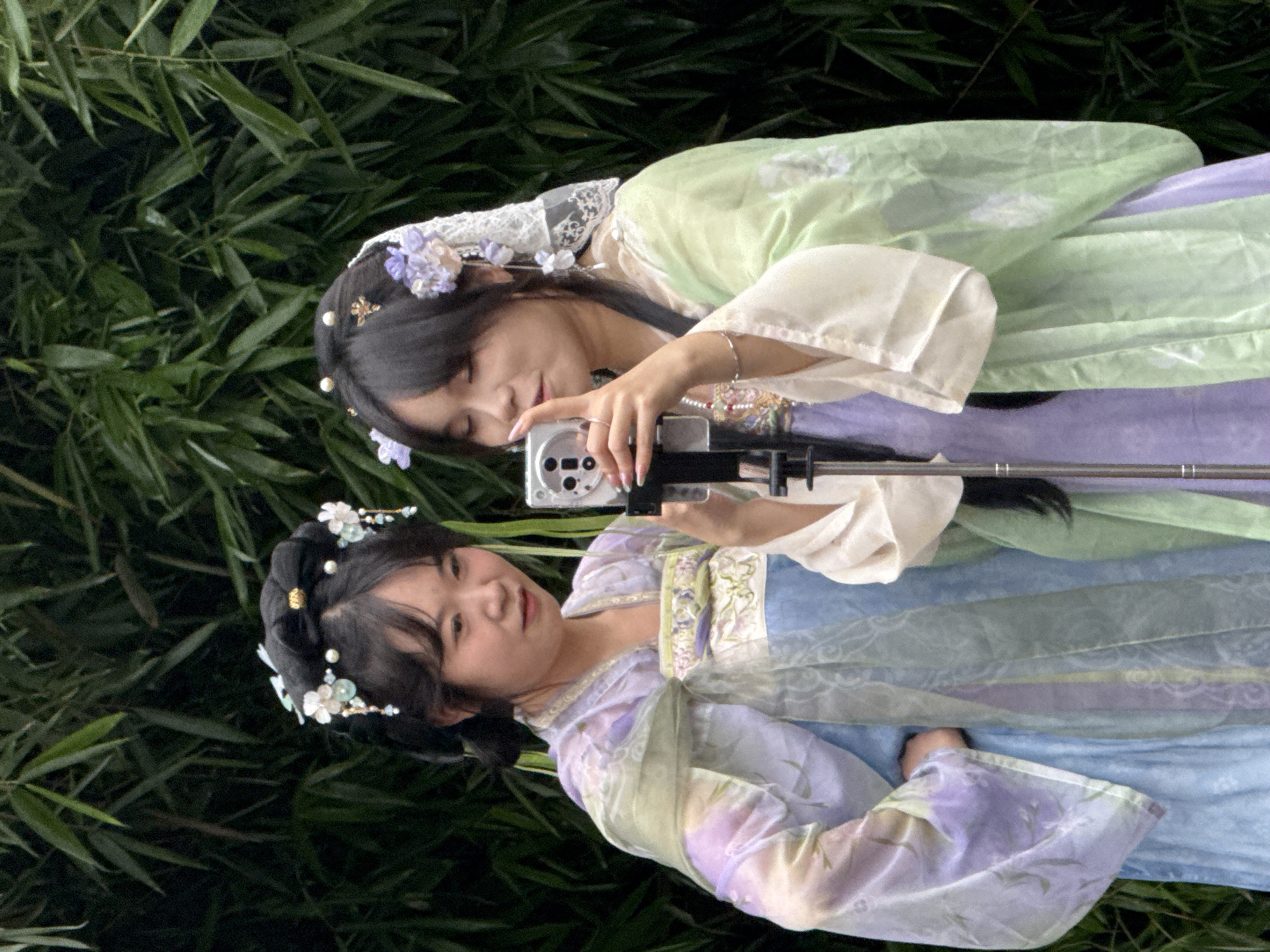 Two young Asian women, dressed in traditional Chinese Hanfu clothing, stand in front of a dense bamboo forest. The woman on the left wears a light purple and pale blue layered garment with sheer, flowing sleeves. She has her hair styled in an intricate updo adorned with white flowers and pearls. The woman on the right is in a light green and pale purple layered Hanfu. Her hair is dark and styled with a lace headpiece and purple flowers. She holds a silver digital camera in her hands, pointing it towards herself as if taking a selfie or a photo of the two of them. Her left hand is raised, touching the camera lens. A silver bracelet is visible on her right wrist. The background consists of dark green bamboo stalks and leaves, creating a natural, serene setting. The lighting suggests it is daytime, possibly overcast, as there are no strong shadows. The image is a medium shot, focusing on the upper bodies and faces of the two women.