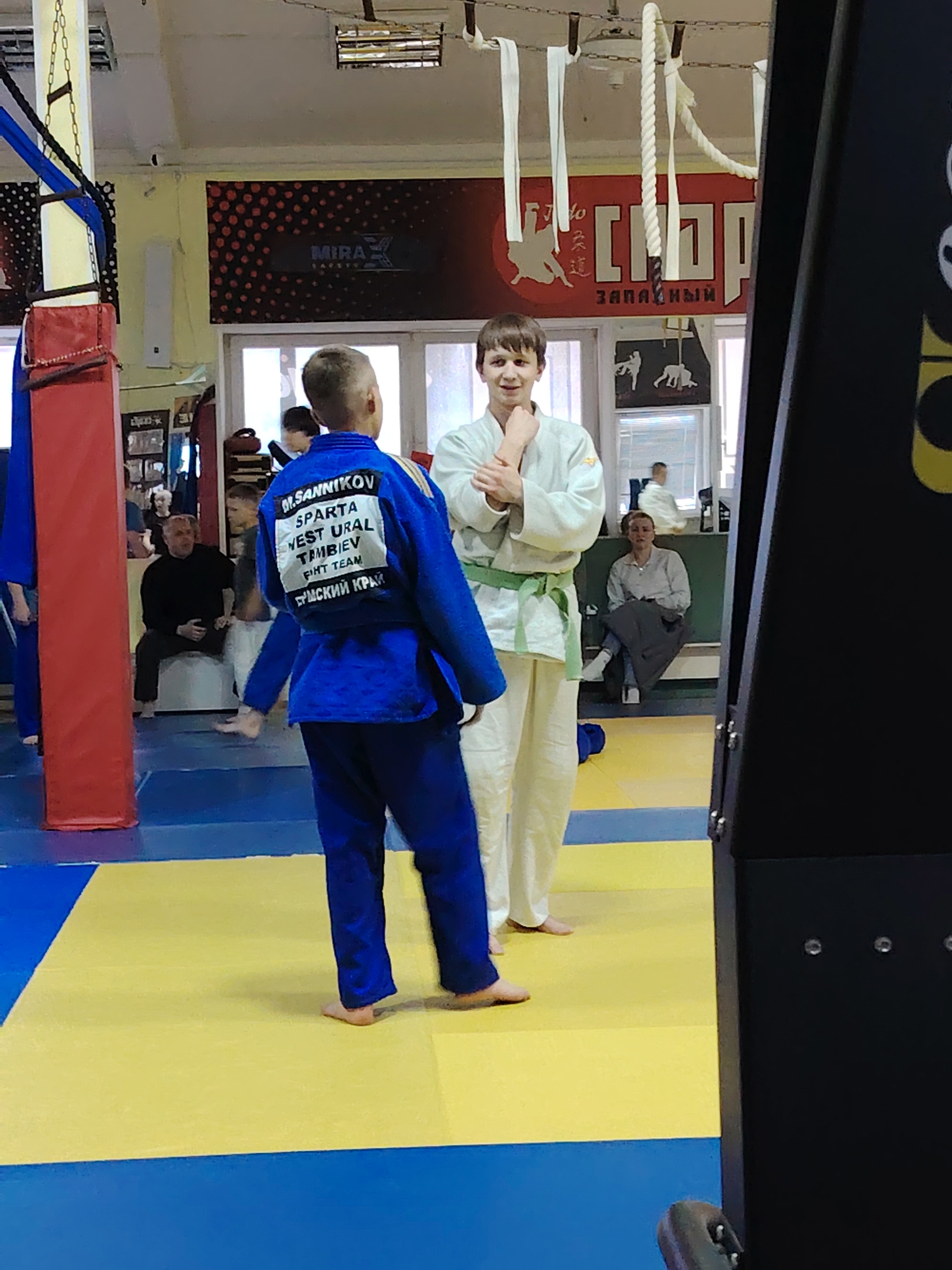The image depicts an indoor judo practice area in Chaikovsky, Russia. Two boys in judo uniforms, one blue and one white with a green belt, stand facing each other on a yellow and blue mat. The boy in blue has text on the back of his uniform identifying "M.SANNIKOV" "SPARTA WEST URAL TAMBIEV FIHT TEAM," and possibly text in Cyrillic. Spectators are seated on benches in the background. Red and black wall signage is visible, including text that reads "MIRA" and Cyrillic characters for "СОРТ" and "ЗАПАДНЫЙ". Gym equipment like a punching bag hangs from the ceiling.