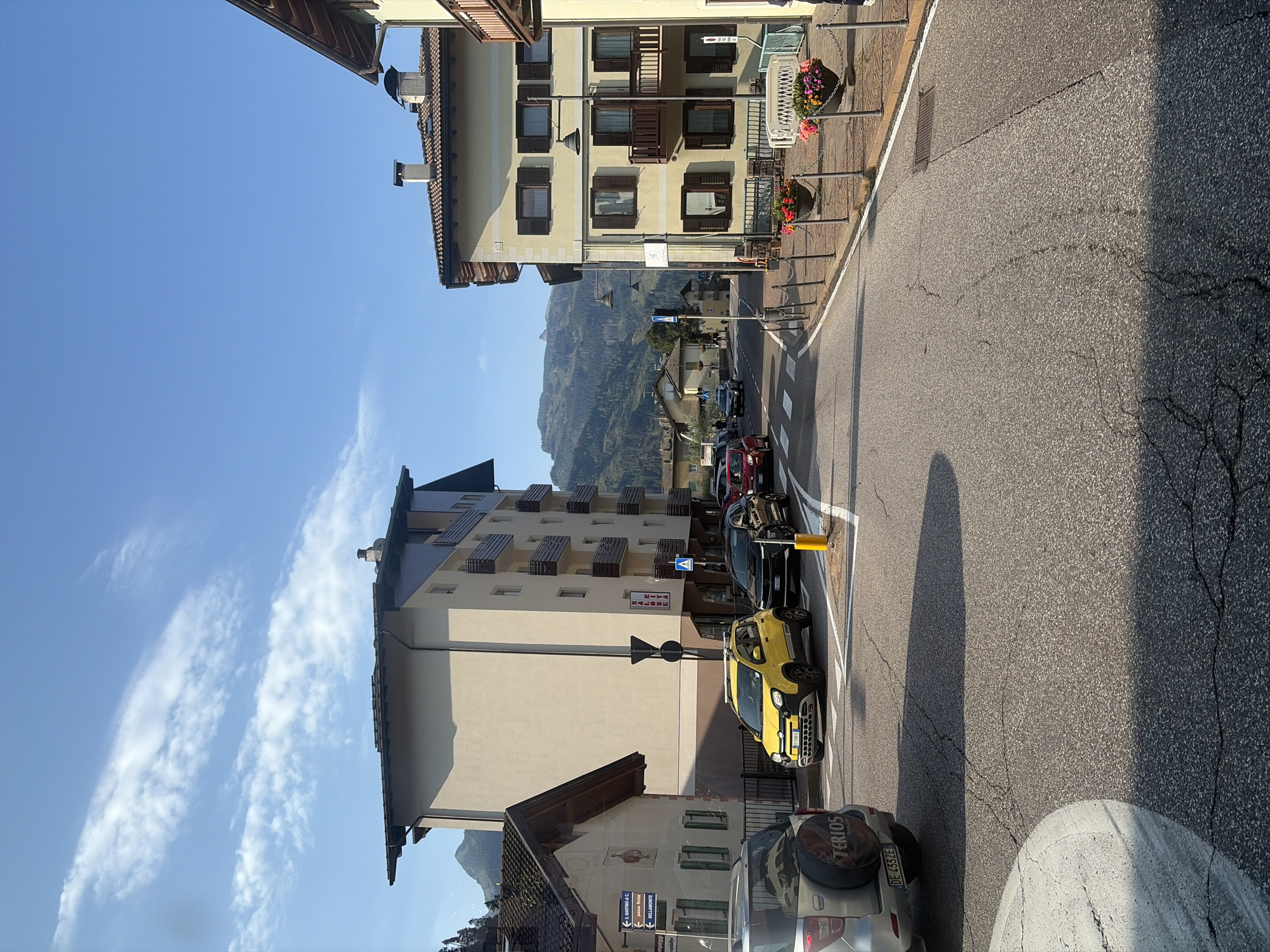 The image depicts a street scene in the town of Predazzo, Italy, likely during the daytime given the clear blue sky with some wispy clouds and the strong sunlight casting shadows. The perspective is from the street level, looking down a paved road that curves to the right.
On the left side of the street, a multi-story building with a light-colored facade and dark-tiled roof stands prominently. This building has balconies with railings and numerous windows. Adjacent to it is a smaller, older building with a darker roof and a decorative facade. Signs are visible on this smaller building, one with arrows indicating directions.
On the right side of the street, another multi-story building with a yellow facade and a dark-tiled roof is visible. This building also features balconies and shutters on its windows. Flower planters with colorful flowers are positioned along the sidewalk in front of this building.
Several cars are present on the road. A silver SUV is parked on the left. A bright yellow small car is stopped at what appears to be a pedestrian crossing or intersection in the middle of the road, along with a black SUV and a red car further down the road. Traffic lights and street lamps are also visible.
In the background, a mountainous landscape with forested slopes and rocky peaks dominates the horizon under the bright sky. The overall impression is of a peaceful, sun-drenched alpine town.