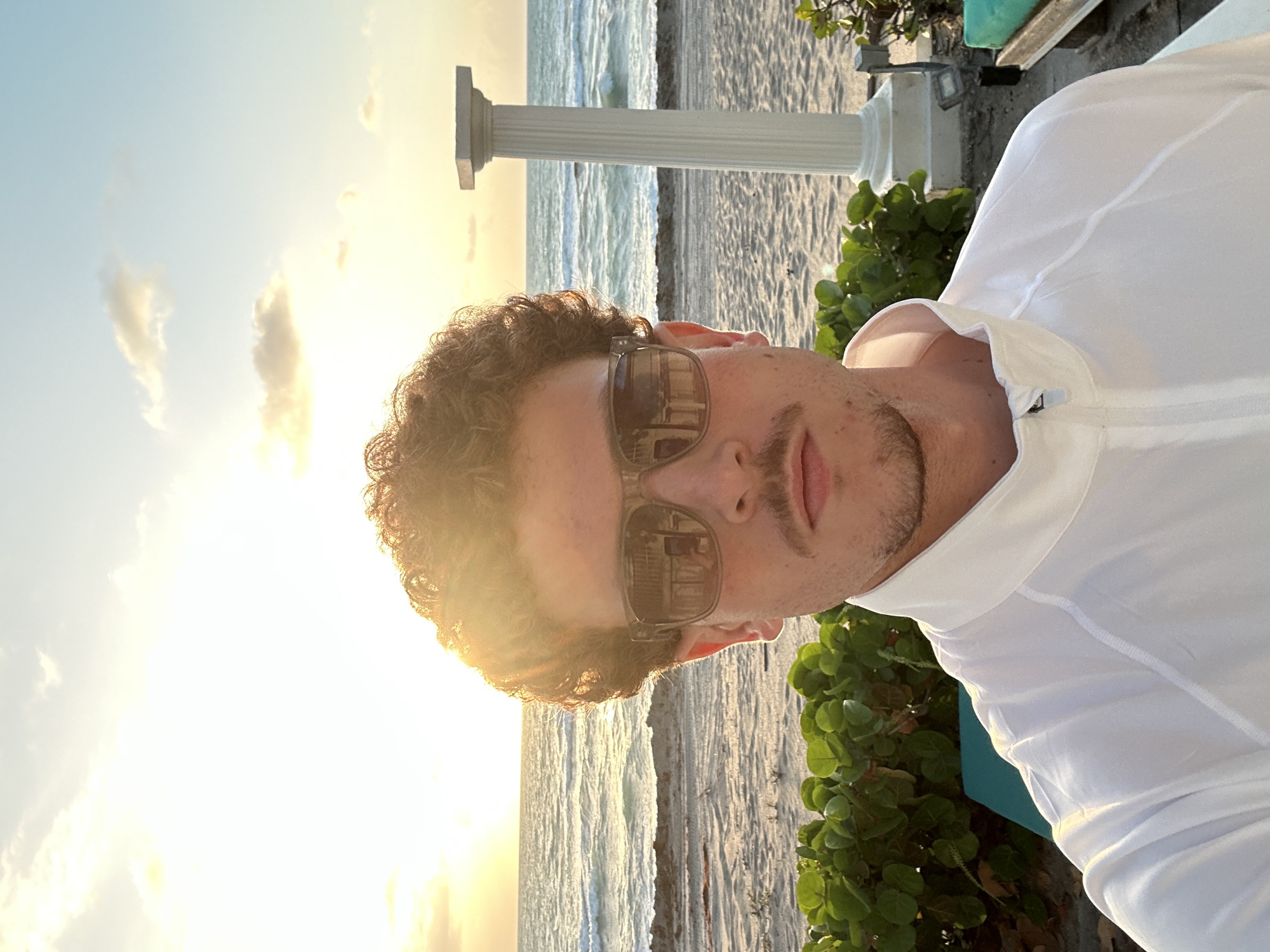 A man with curly brown hair and sunglasses is standing on a beach. He is wearing a white jacket. Behind him is the ocean and a sandy beach. A white column is visible in the background, along with green foliage. The location is Hallandale Beach, United States of America. The time of day appears to be sunset or sunrise, based on the bright sky.