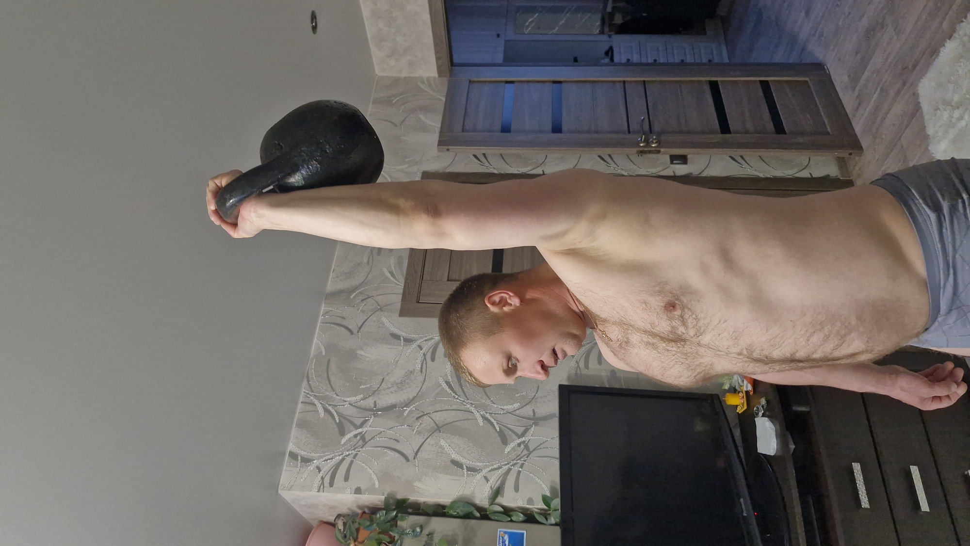 A shirtless man, fair-skinned with short light brown hair and visible chest and abdominal hair, wearing grey boxer briefs or shorts, is captured mid-exercise in what appears to be a residential room in Otchor, Russia. He is performing a strength training or stability exercise, holding a large black kettlebell overhead with his right arm fully extended. His left hand also appears to be gripping the kettlebell for support. His expression conveys focus and effort.
The indoor setting features a wall adorned with grey patterned wallpaper, characterized by swirling, possibly floral or abstract, designs. To the man's right, a dark wooden door with paneling is visible, along with glimpses of a light-colored wooden floor. To his left, a dark wooden dresser or cabinet holds a black flat-screen television and a green potted plant. A white, possibly furry, rug is partially visible on the floor to the far right. The room is illuminated by artificial lighting, as no natural light sources are discernible, suggesting the photo could have been taken at any time of day. No text is visible in the image.