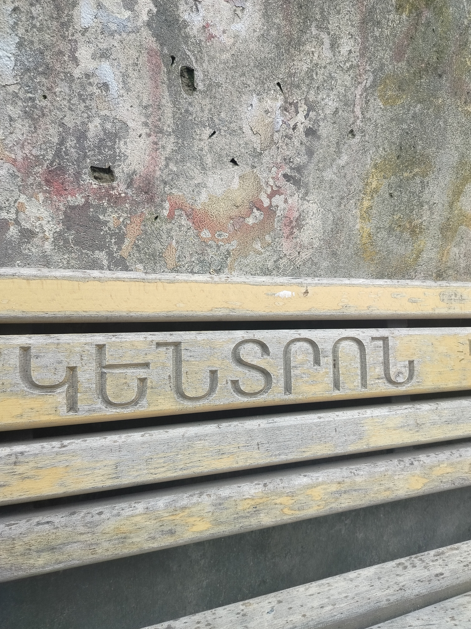 The image shows a close-up of a wooden bench with the Armenian word "ԿԵՆՏՐՈՆ" carved into one of its slats. This word translates to "Center" in English. The bench is weathered, with peeling paint, suggesting age and exposure to the elements. The background consists of a textured, decaying wall, with patches of discoloration and visible repairs. The location is Yerevan, Armenia. The focus is on the bench and the carved word, indicating it's a significant detail within the scene. There are no people visible.
