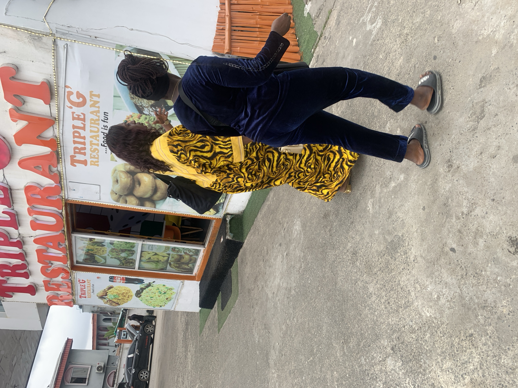 The image shows the exterior of a restaurant named "TRIPLE G RESTAURANT" located in Lagos, Nigeria. Two women are walking past the restaurant entrance. The woman on the left is wearing a bright yellow dress with a black abstract pattern, and the woman on the right is wearing a navy blue velour tracksuit. The restaurant has a glass storefront displaying pictures of food items. A black Toyota sedan with its hood up is parked in the background, with a person attending to it. The ground is paved with asphalt and there are some patches of artificial green turf near the restaurant entrance.