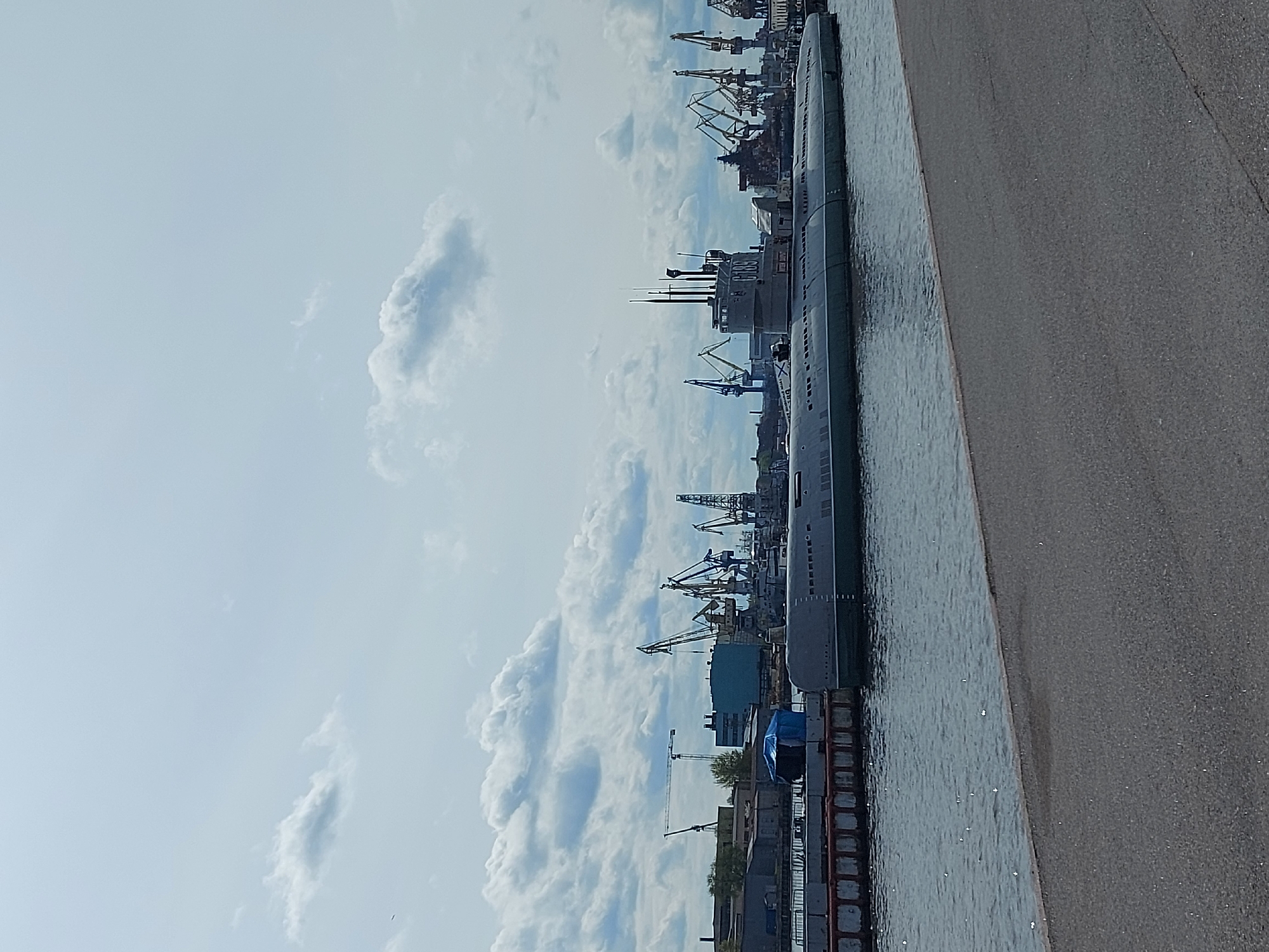 The image shows a submarine docked in Saint Petersburg, Russia. The submarine is gray and long, situated in the water alongside a concrete embankment. In the background, the cityscape is dominated by numerous cranes, suggesting a busy port or industrial area. The sky is cloudy, with patches of blue visible. No people are visible in the image.