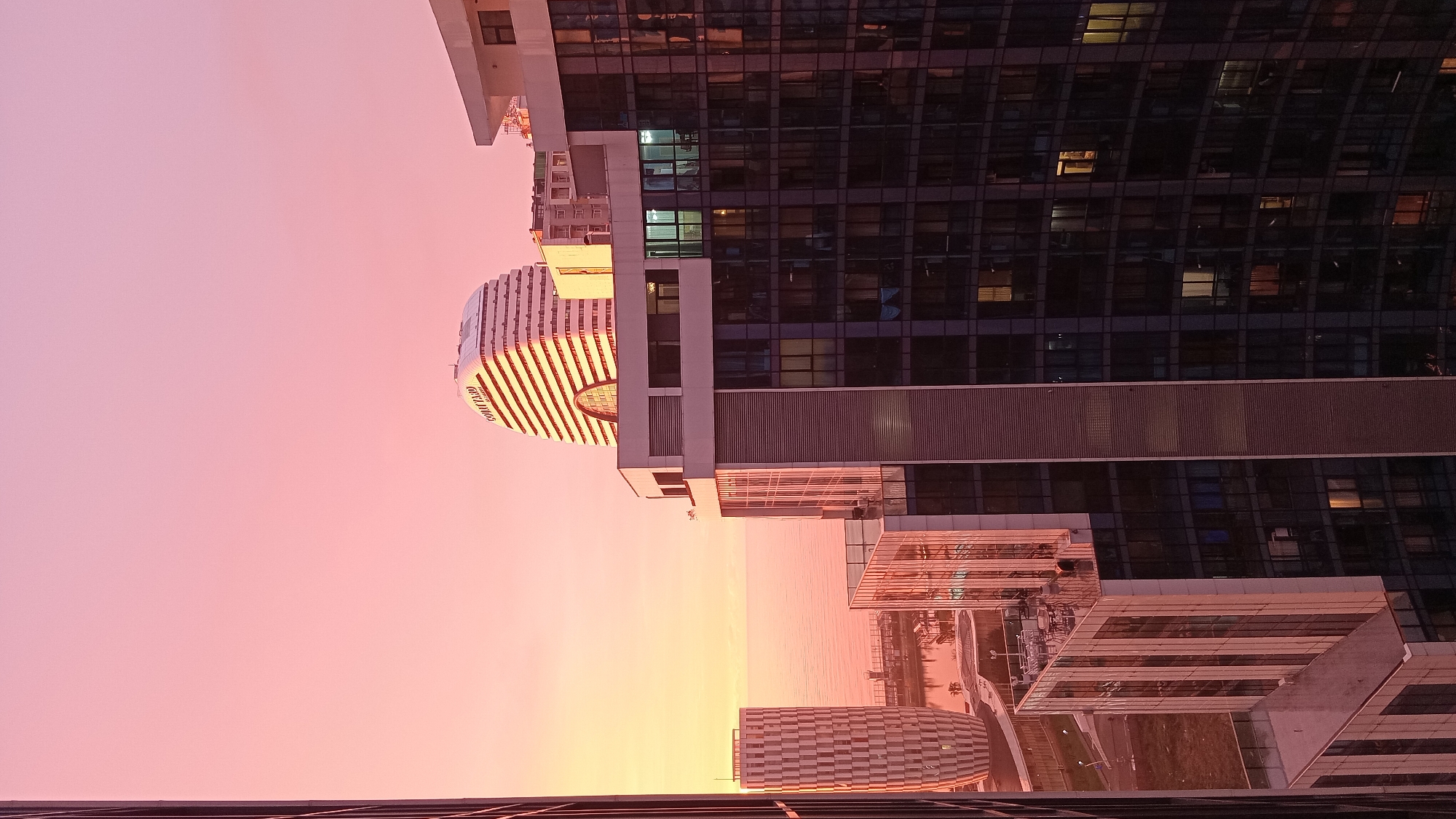 The image displays an elevated urban landscape view of Batumi, Georgia, during either sunrise or sunset. The sky exhibits a color gradient from pink at the top to orange and yellow hues near the horizon, which meets the Black Sea.
In the mid-ground, the Courtyard by Marriott Batumi hotel is prominently featured, identifiable by its white, curved, and horizontally ribbed facade with "COURTYARD" lettering visible on its upper portion. To the left of this structure, a tall, cylindrical building with a distinctive geometric patterned exterior stands near the waterfront.
The immediate foreground is dominated by multiple modern high-rise buildings characterized by dark, reflective glass exteriors. These structures partially frame the scene; a building on the right side shows several illuminated interior windows. A segment of the city's ground level, including paved areas and waterfront infrastructure, is visible between the foreground buildings and the sea. No individuals, specific actions, or interactions are discernible within the frame.