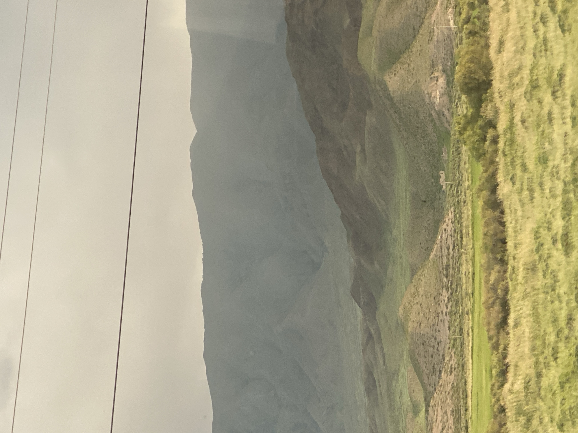 The image depicts a wide, expansive landscape with rolling hills and mountains under a hazy sky. Three thin, dark horizontal lines, possibly power lines or telephone wires, stretch across the upper portion of the frame, obscuring a small part of the sky and the distant mountain range. The mountains in the background are layered, with the farthest peaks appearing muted and gray due to atmospheric haze. The middle ground features undulating hills covered in a mix of dry brush and green vegetation. In the foreground, a field of tall, dry grass dominates, with a patch of vibrant green grass to the left and a line of green bushes and trees further back. The scene appears to be an outdoor, natural environment, with no visible people or specific man-made structures apart from the overhead wires and a faint white pole in the middle ground. The overall impression is one of a vast, somewhat arid, and undeveloped natural landscape. The location is identified as Kok-Moinok - 1, Kyrgyzstan.