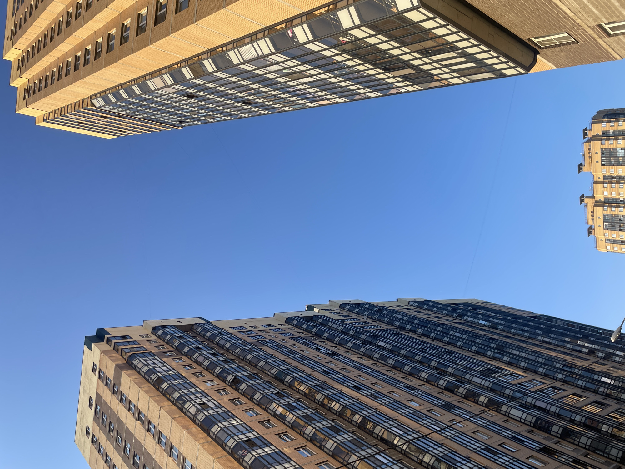 The image captures a clear, cloudless blue sky above a cluster of tall residential buildings in Pargolovo, Russia. Two prominent high-rise apartment buildings dominate the frame, one on the left and one on the right, both angled upwards towards the sky. The building on the left, with a facade of light brown brick and numerous dark-framed windows, has a section of balconies with glass railings. The building on the right, also constructed of light brown brick, features a repeating pattern of windows and a striking diagonal element of glazed balconies. In the distance, towards the bottom center of the image, a smaller residential building is visible with a more complex, stepped roofline. The perspective is from a low angle, looking up at the structures against the expansive sky. No people are discernible within the image.