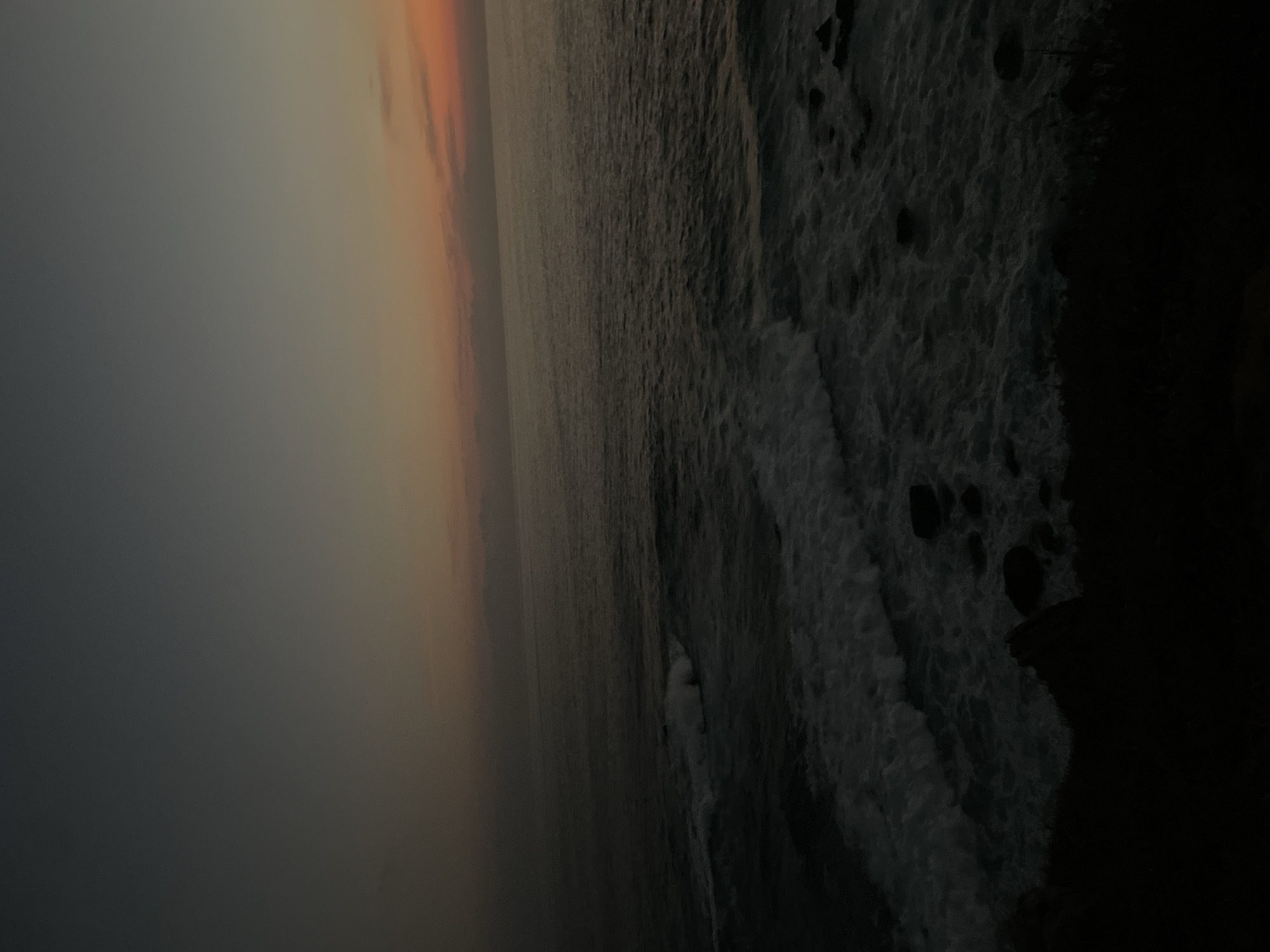 The image depicts a coastline at sunset in Puerto Escondido, Mexico. The sky transitions from a dark gray-blue to orange, suggesting either sunrise or sunset. The ocean is a dark expanse, with visible waves crashing against a rocky shoreline to the right of the frame. The shoreline itself appears rugged, with dark rocks and vegetation merging into shadows. There are no visible people or distinct objects other than the natural elements.