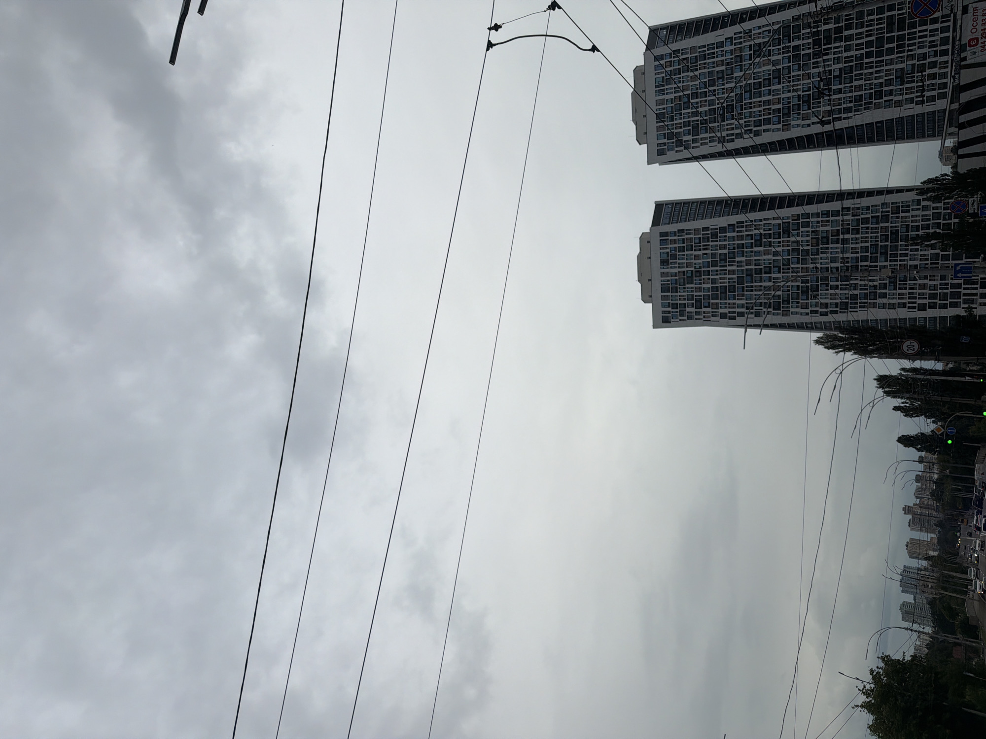 The image shows a city skyline in Kyiv, Ukraine, under a cloudy sky. In the foreground, there are multiple overhead power lines and some trees on the left and right sides of the frame. In the middle ground, two tall, modern residential buildings with many windows are visible. In the background, a cityscape with more buildings and streetlights can be seen. The street below is lined with cars, indicating a busy urban environment. The overall mood of the image is overcast and subdued due to the cloudy weather.