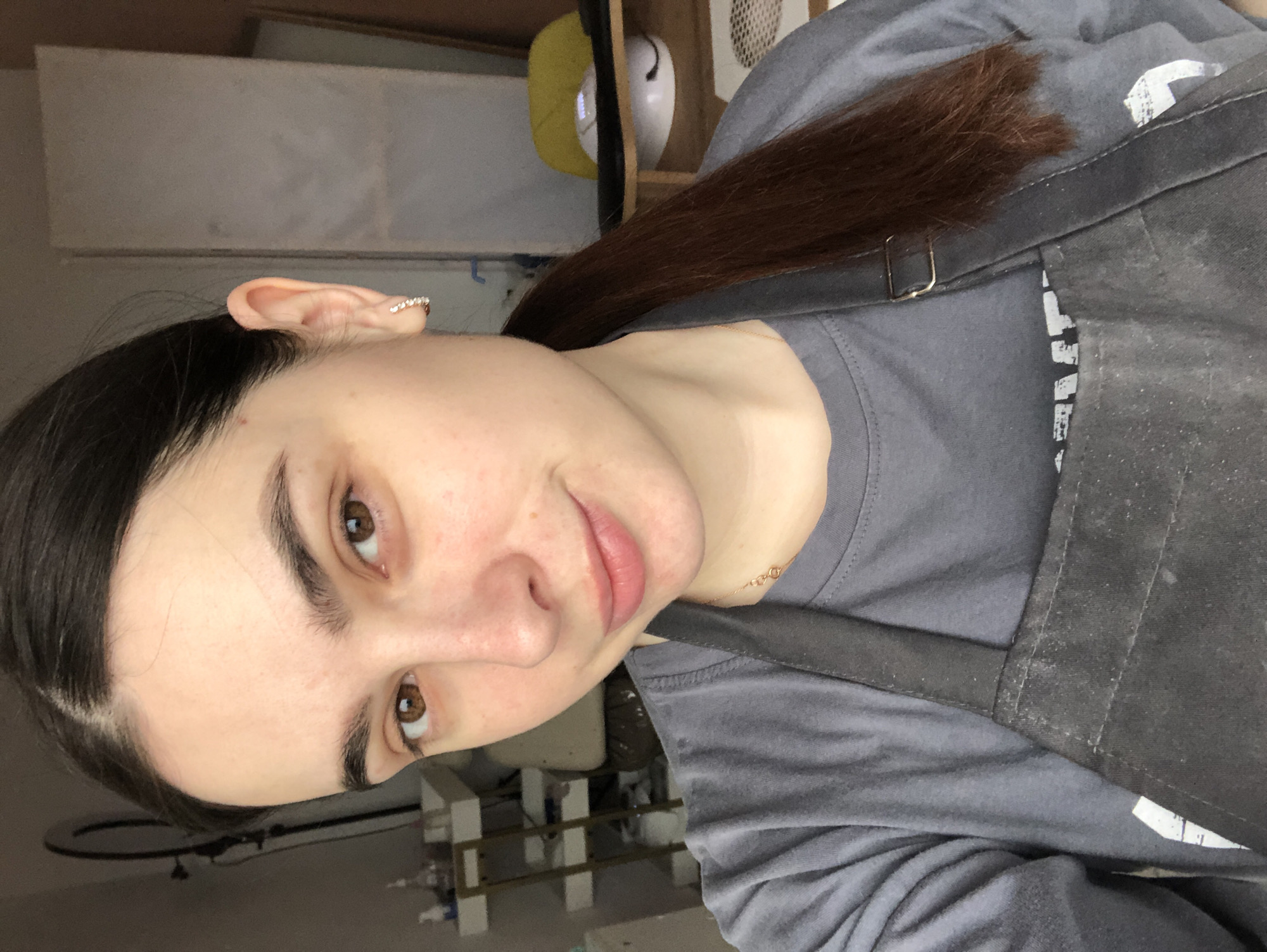 The image is an indoor, head-and-shoulders portrait of a young woman. She has light skin, dark brown hair pulled back into a ponytail, and warm brown eyes. She looks directly at the camera with a neutral to slightly smiling expression. She wears a plain grey short-sleeved t-shirt and a dark grey or black apron, visible over her right shoulder with a metallic buckle. A thin gold chain is visible around her neck, and a delicate ear cuff with small clear stones is on her right ear. The apron appears to have some light dust or residue on its surface.
The background suggests an indoor workshop or studio setting. Behind her head, a light-colored wall or shelving unit is present. To her right, a yellow object, possibly a cap, and a white object, perhaps a mask or small device, are visible on a shelf. A black ring light stand is partially visible in the lower left portion of the background. Further down, some white structures, possibly shelves or equipment frames, can be seen. A partial, illegible white logo or text is visible on her grey t-shirt, beneath the apron strap. No specific time of day or weather can be discerned. The user provided the location context as Chaikovsky, Russia, but this is not visually confirmed within the image.