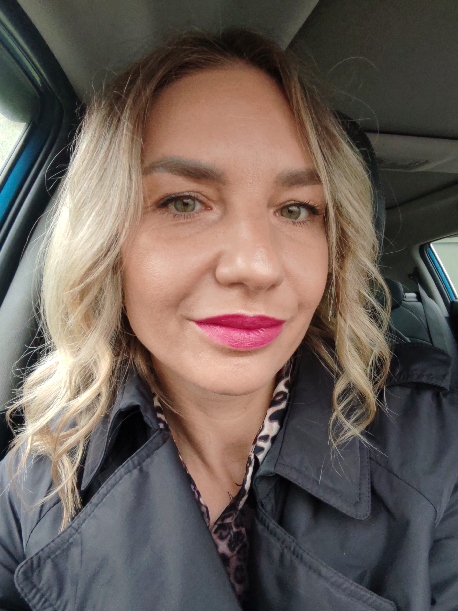 A close-up, eye-level shot captures a woman seated inside what appears to be a car. She has blonde, wavy hair that falls around her face and shoulders. Her eyes are a striking green, and she is wearing bright pink lipstick. Her complexion is smooth, and a faint smile plays on her lips as she looks directly into the camera.
She is dressed in a dark, possibly black, trench coat with a prominent collar. Beneath the coat, a garment or scarf with a leopard or animal print pattern is visible. The background shows the interior of a vehicle, with a blue trim piece visible on the left, suggesting a car door frame. Black car seats are partially visible behind her, and the light grey ceiling of the car is above. The lighting suggests it is daytime, possibly overcast or with diffused light entering the vehicle. There is no visible text in the image.