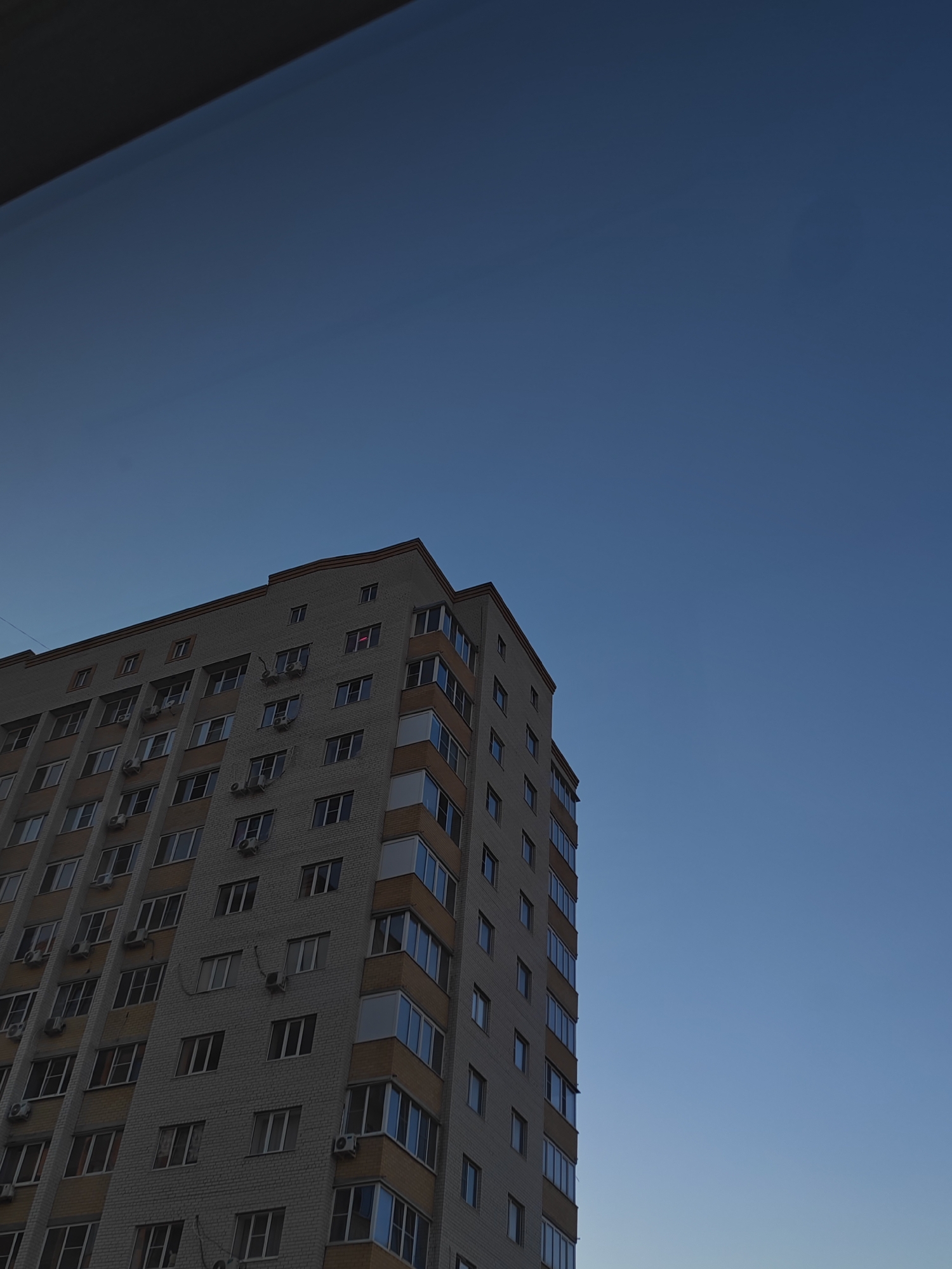 An exterior, low-angle, full shot captures the corner of a light-colored brick apartment building against a deep blue sky. The building has many windows, some open, and several air conditioning units are mounted on the exterior walls. Orange trim runs along the balconies and some window frames. The top edge of the building is visible, as is a dark overhang or structure in the upper left corner of the image. The sky is clear, suggesting it is either early morning or late evening. No people are visible in the image. This scene is consistent with the architecture found in residential areas of Bataysk, Russia.