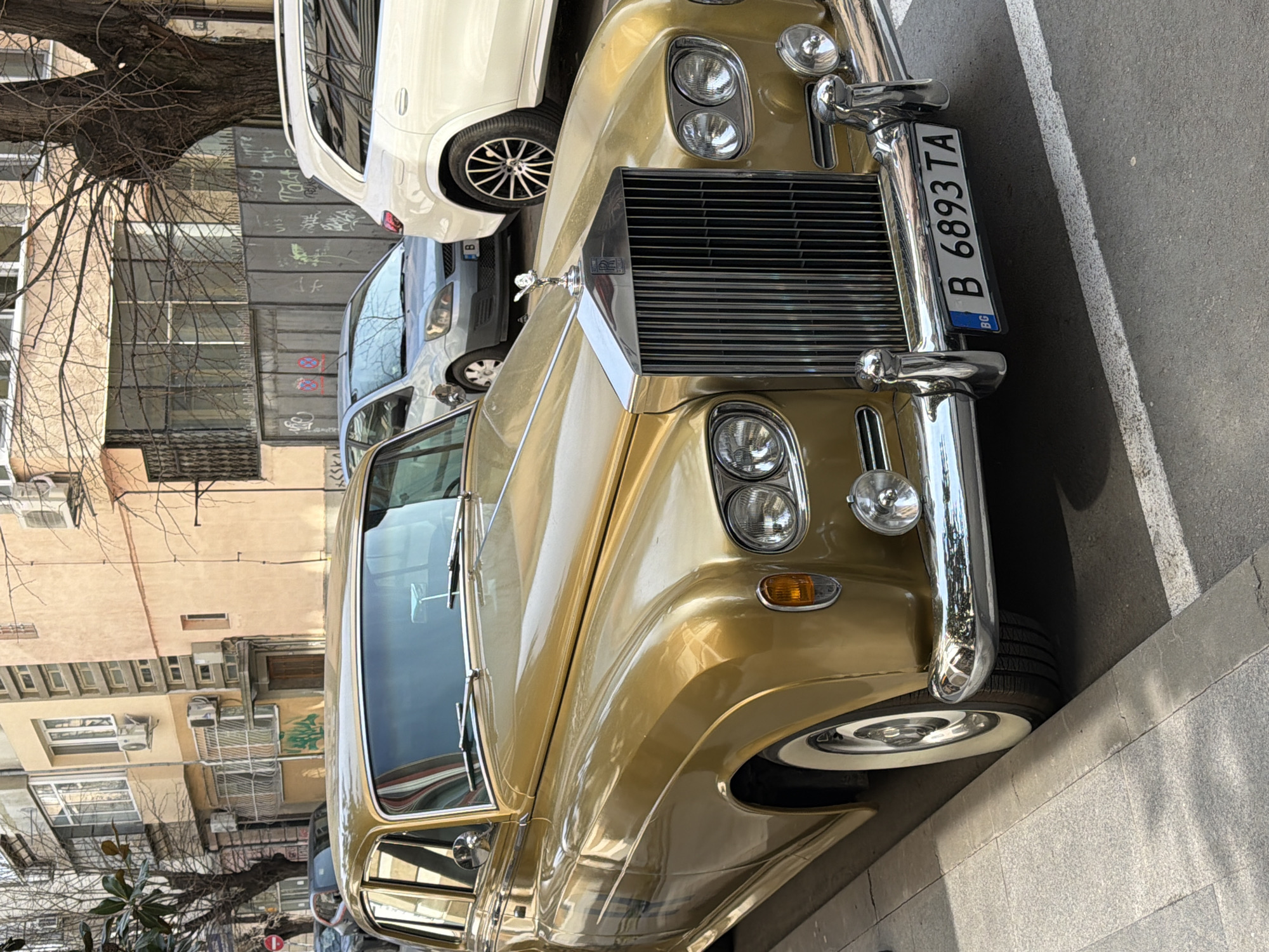 An urban street scene in Varna, Bulgaria, depicts a vintage metallic gold Rolls-Royce sedan parked parallel to a light grey tiled sidewalk. The Rolls-Royce, facing towards the viewer, features a prominent chrome grille with vertical slats, a "Spirit of Ecstasy" hood ornament, and a "Rolls-Royce" emblem. It has dual circular headlights on each side, amber turn signal indicators, a chrome bumper with two additional auxiliary lights, and white-wall tires. The Bulgarian front license plate displays "B 6893 TA," with the "B" prefix indicating registration in Sofia.
Behind the Rolls-Royce, a silver/grey hatchback and a white SUV with multi-spoke dark alloy wheels are also parked on the street. The background consists of multi-story residential or mixed-use buildings with facades in shades of beige and light orange, featuring multiple windows, balconies, and externally mounted air conditioning units. Bare deciduous trees line the street, indicating a cooler season. A metal fence or gate with graffiti is visible further back, along with a partially visible red circular "No Entry" sign on the far left. The scene is illuminated by clear daylight, casting distinct shadows. No individuals are present in the image.