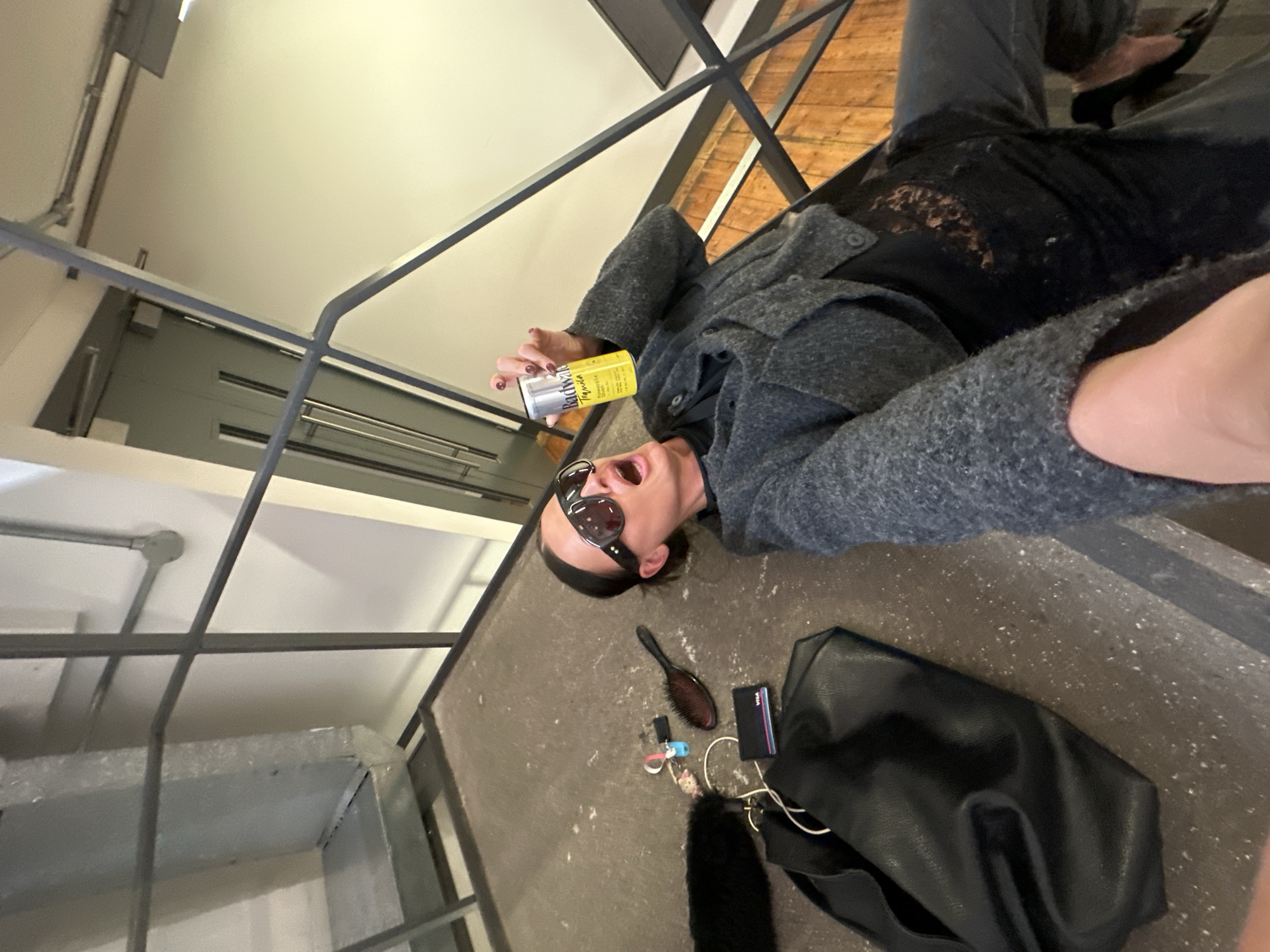 An individual is depicted lying on their back on a grey, speckled concrete floor within what appears to be an indoor setting. The image is taken from an overhead perspective and rotated sideways, showing the person in the lower half of the frame.
The person is wearing dark sunglasses, a textured grey jacket with large buttons, and a black top featuring lace detailing across the chest. Their dark hair is pulled back, and their mouth is wide open, suggesting an expression of excitement, shouting, or laughter. In their right hand, they hold a yellow and silver can, on which the text "Bachman's Tequila" is clearly visible, along with other smaller, unreadable text. Their fingernails are painted red.
Scattered on the concrete floor near the person's head are several personal items: a black oval-shaped hairbrush, a black wallet with a "VISA" card peeking out, a black leather-like handbag, and a small cluster of items resembling keys or accessories, including pink and blue elements. A black furry item is also near the bag.
In the background, behind metal railings, there is a section of wooden flooring and a light-colored wall. Above and to the side, more industrial-style metal structures and light-colored ceilings/walls are visible. The setting suggests a modern or industrial interior, possibly a building in London, UK.