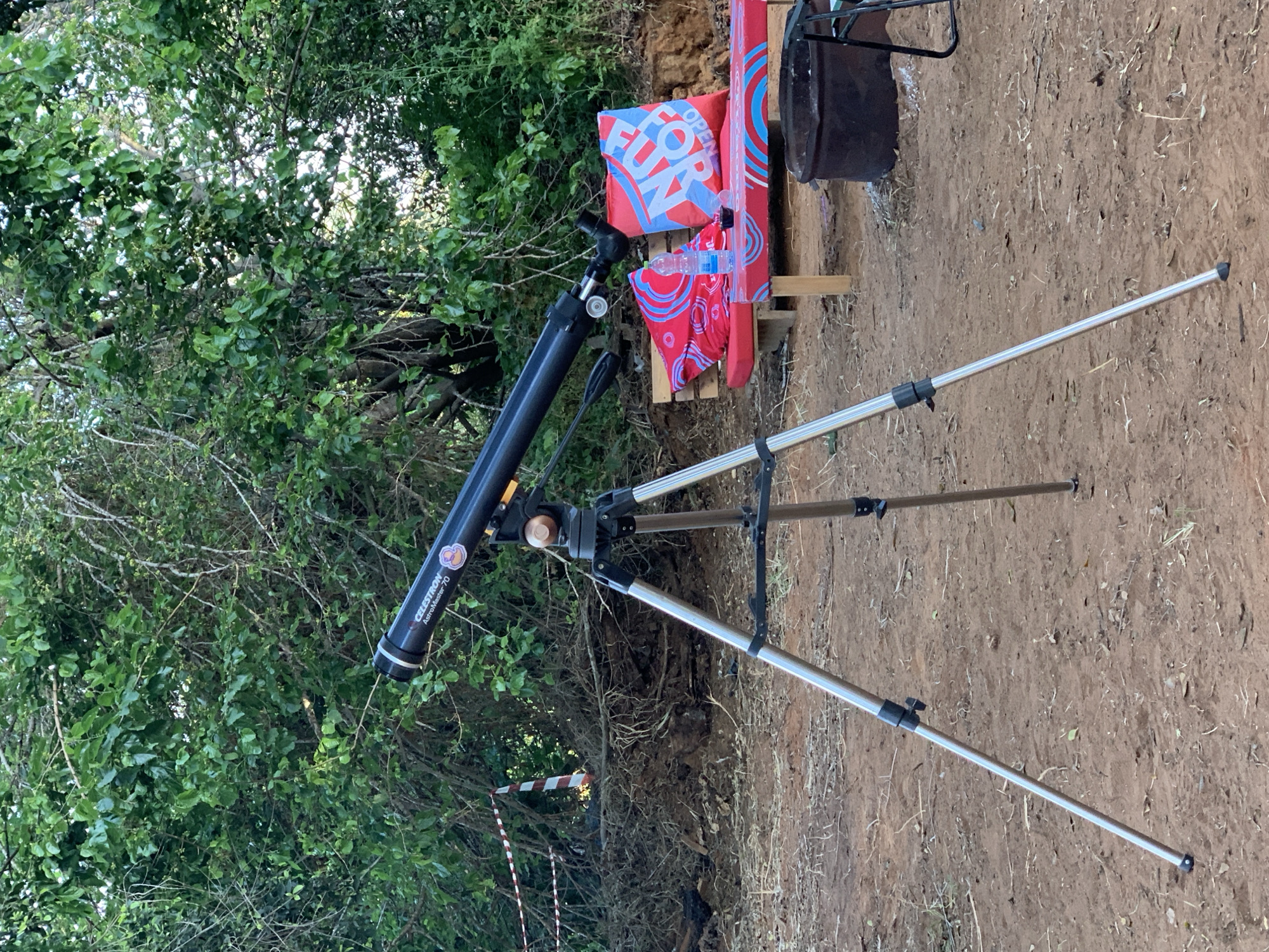 The image shows a Celestron AstroMaster 70 telescope on a tripod, positioned outdoors near a bush and an "OPEN FOR FUN" sign with red and blue patterns. The setting appears to be in a natural area with dirt ground, possibly in the vicinity of Gan Sorek, Israel, given the provided location. There is a trash can behind the sign. A red and white striped barrier tape is visible near the bush and telescope legs. There are no people visible in the scene.