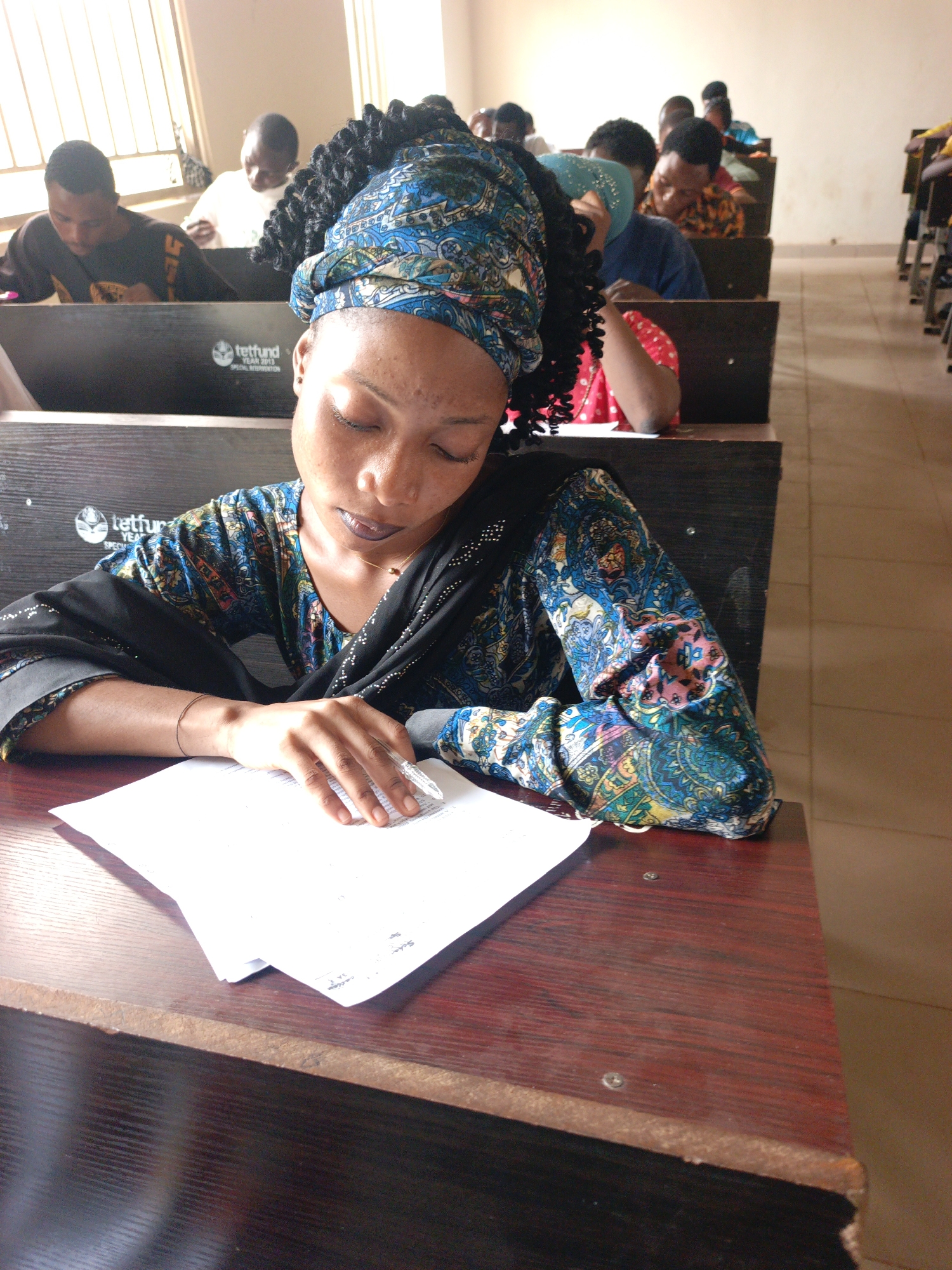 The image depicts a woman in a classroom setting in Akwanga, Nigeria. She is seated at a wooden desk with papers in front of her, apparently reading or writing. She wears a colorful, patterned top and a matching headwrap. Other people are seated at desks behind her, also appearing to be engaged in study or work. The room has rows of desks, indicating a classroom or lecture hall environment. "Tetfund" is printed on the front of the desks. The overall impression is one of a classroom or learning environment.