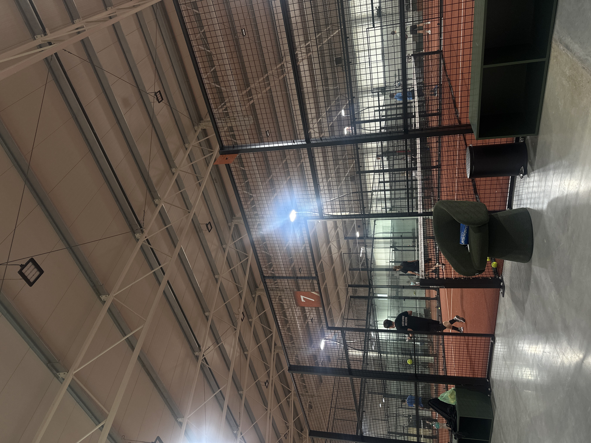 This image captures an indoor padel tennis facility, presented in a portrait orientation where the original capture was rotated 90 degrees clockwise. The scene features multiple padel courts, characterized by their reddish-brown playing surfaces, transparent glass back walls, and black metal mesh fencing that encloses the playing areas.
The facility boasts a high, arched ceiling constructed from white corrugated metal panels supported by a network of white trusses and structural beams. Numerous bright LED light fixtures are suspended from the ceiling, providing ample illumination for the courts.
Several individuals are visible on the courts, actively engaged in playing or practicing padel tennis. In the immediate foreground, next to one of the courts, there is a green, upholstered armchair with "Babolat COURT" visible on its side, a black cylindrical trash can, and a dark green, open-shelving unit. Yellow padel balls are scattered across the court surfaces.
A player in a black t-shirt is prominent in the mid-ground, with the partial text "BLACK HORSE RANCH & COUNTRY CLUB" visible on his back. A large, rectangular sign, orange-brown in color and bearing the number "7", hangs from the ceiling structure, likely indicating the court number. The overall impression is that of a spacious, well-maintained, and modern indoor sports complex.