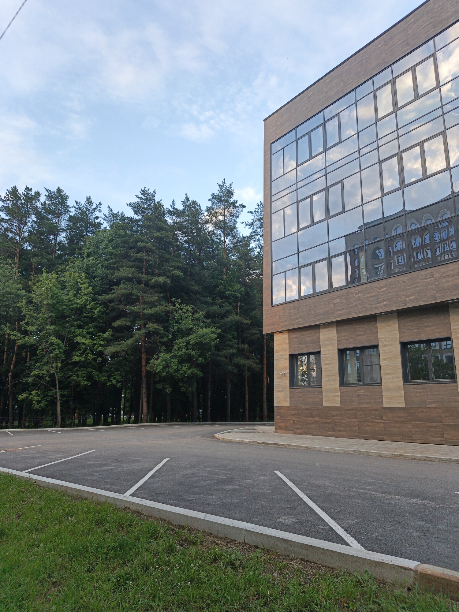 The image depicts a modern building with a facade of glass and wood-like panels, reflecting the sky and clouds. The building has multiple rows of rectangular windows. To the left of the building is a dense forest of tall pine trees and other deciduous trees. In the foreground, there is an asphalt parking lot with white parking lines. Green grass borders the parking lot. The sky is partly cloudy and appears to be daytime. This scene is located in Ufa, Russia, as confirmed by similar images found online and the architectural style consistent with buildings in that region.