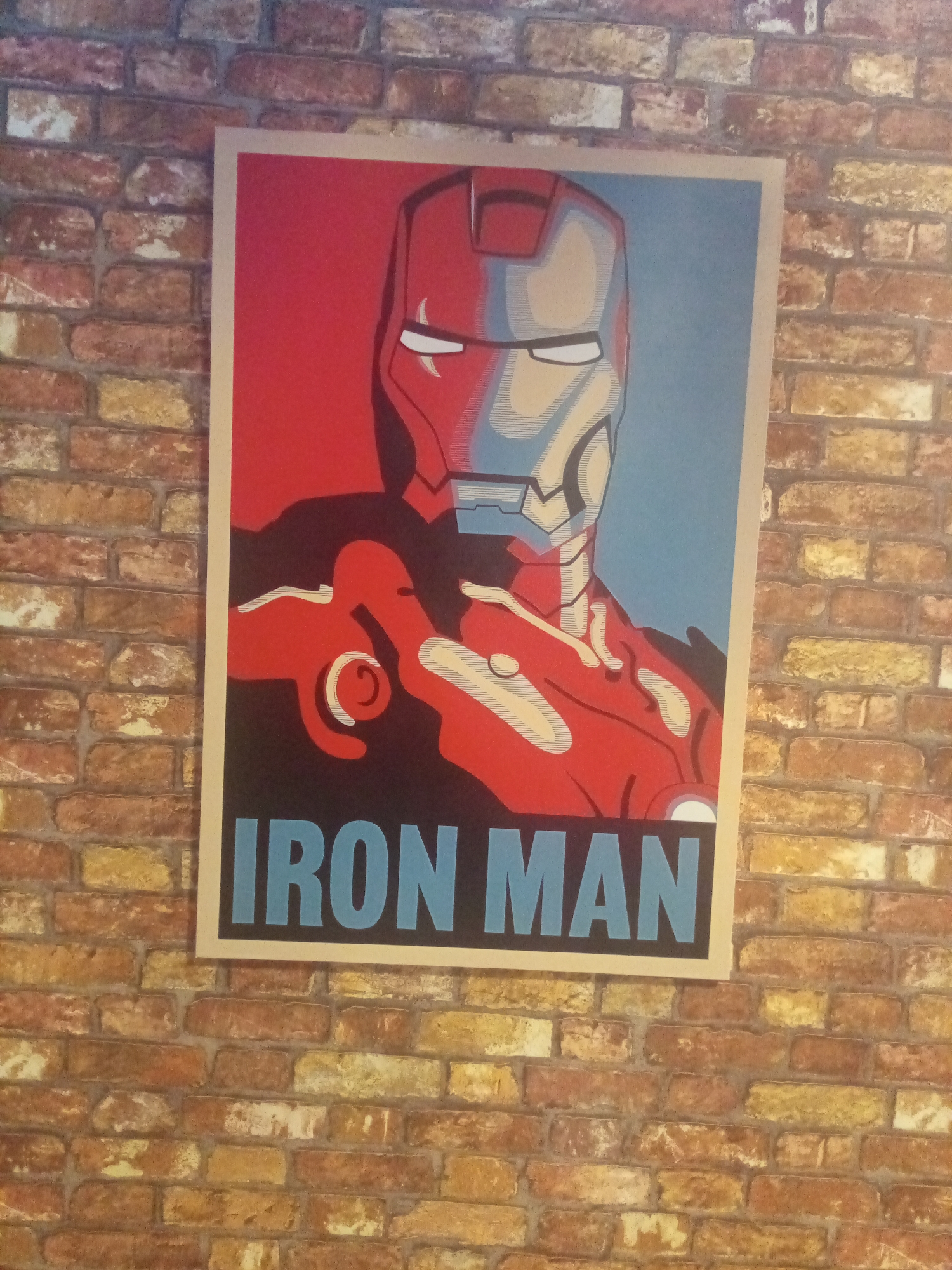A framed poster of Iron Man is displayed on a brick wall. The poster features a stylized portrait of Iron Man's helmet and upper torso, split into red and blue sections. The text "IRON MAN" is printed in large, light blue letters across the bottom of the poster. The brick wall has a mottled appearance with variations in color, ranging from reddish-brown to yellow and off-white. The lighting suggests an indoor environment, possibly a cafe, bar, or entertainment venue. The location is specified as Porlamar, Venezuela.