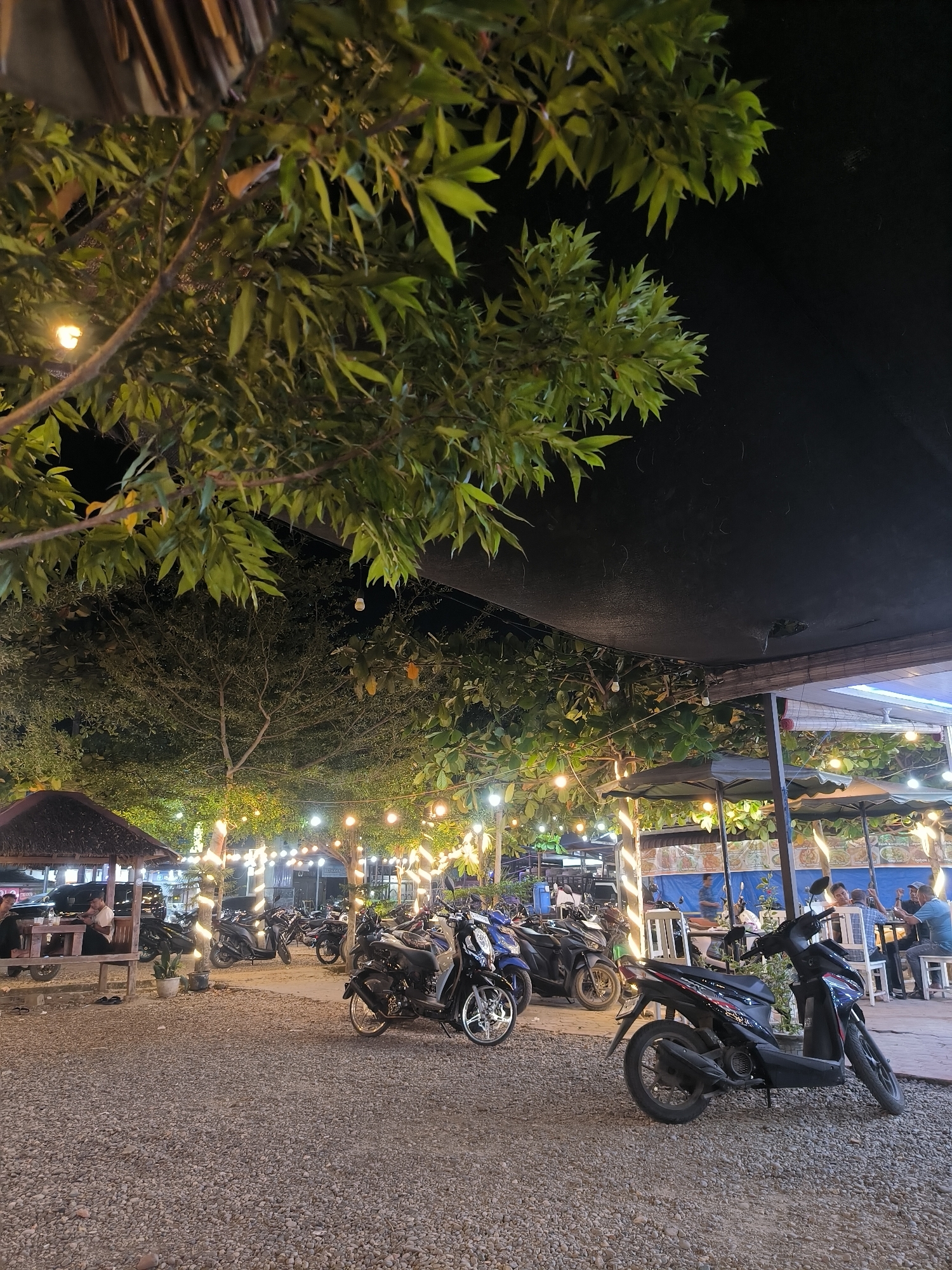 This outdoor scene in Titibaro, Indonesia, shows a gravel lot filled with parked motorcycles. A thatched-roof gazebo sits to the left, occupied by people. Strings of lights illuminate the trees overhead, creating a festive atmosphere. In the background, people are seated at tables under canopies.