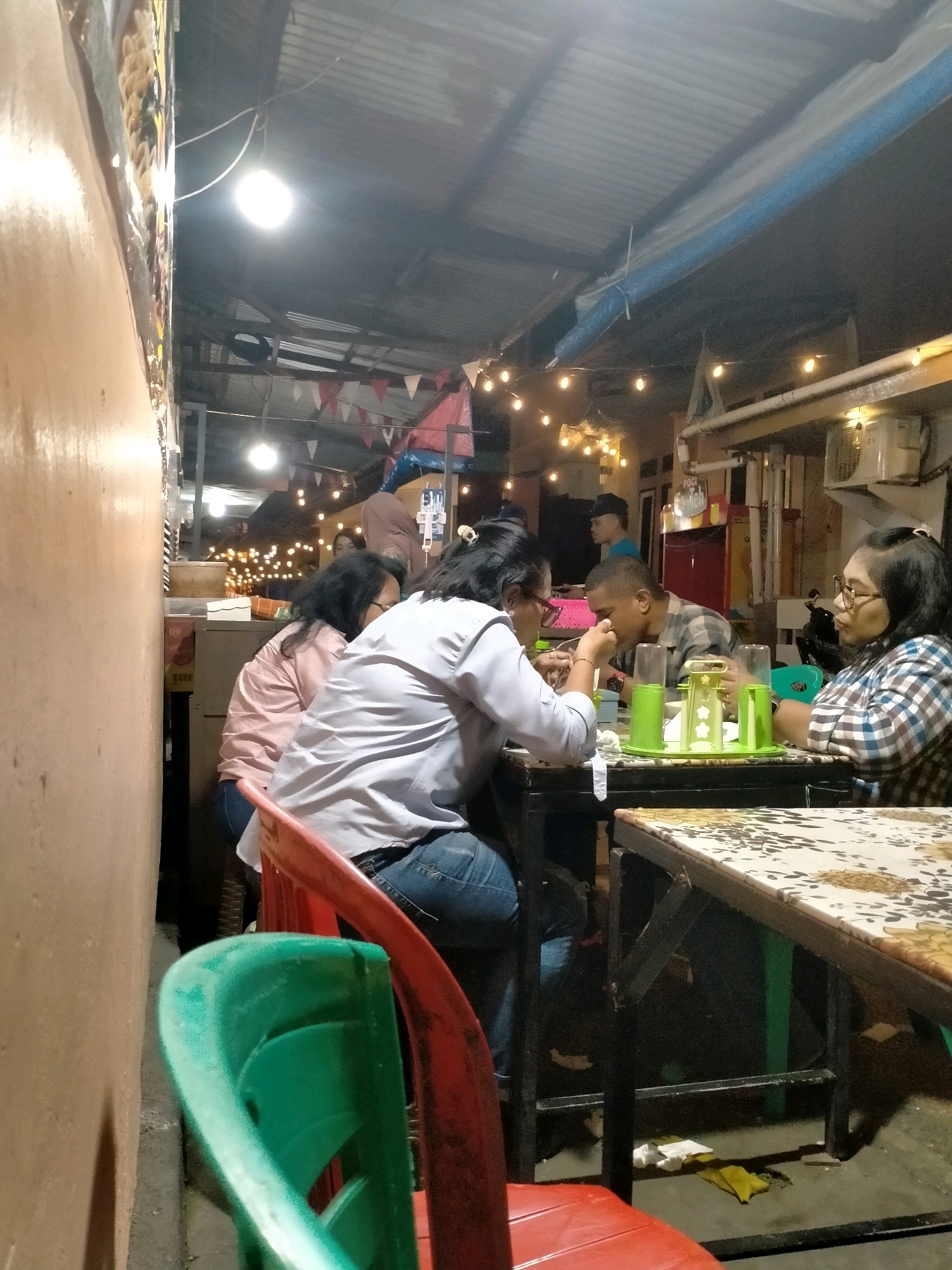 The image depicts a dimly lit outdoor dining area in Ambon, Indonesia. The scene features several people seated at tables, engaged in eating and conversation. The environment suggests a casual, perhaps street-side eatery, with a corrugated metal roof overhead and string lights illuminating the space. The overall atmosphere is relaxed, indicating a common social gathering.