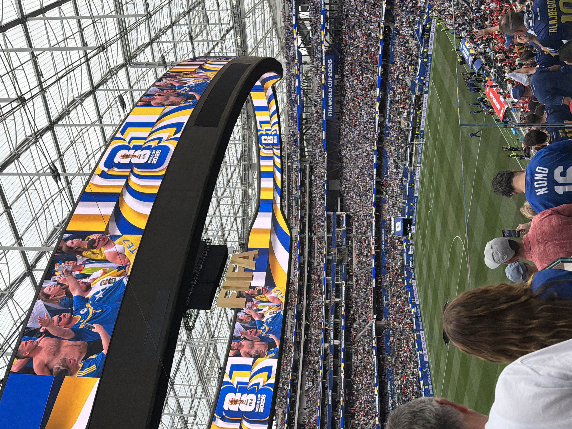 The image shows the interior of SoFi Stadium in Inglewood, California, during an event related to the FIFA World Cup 2026. A massive, curved video screen displays "FIFA World Cup 2026" with the FIFA trophy and colorful graphics. Below the screen, a large crowd of spectators fills the stadium seats, most wearing blue and white. The playing field, a vibrant green soccer pitch, is visible in the foreground, with several individuals on the field, including what appear to be players in blue uniforms and staff members in yellow vests. Some spectators in the lower seating areas are also visible, with one individual holding up a phone to record the event. The stadium's distinctive roof structure is visible overhead.