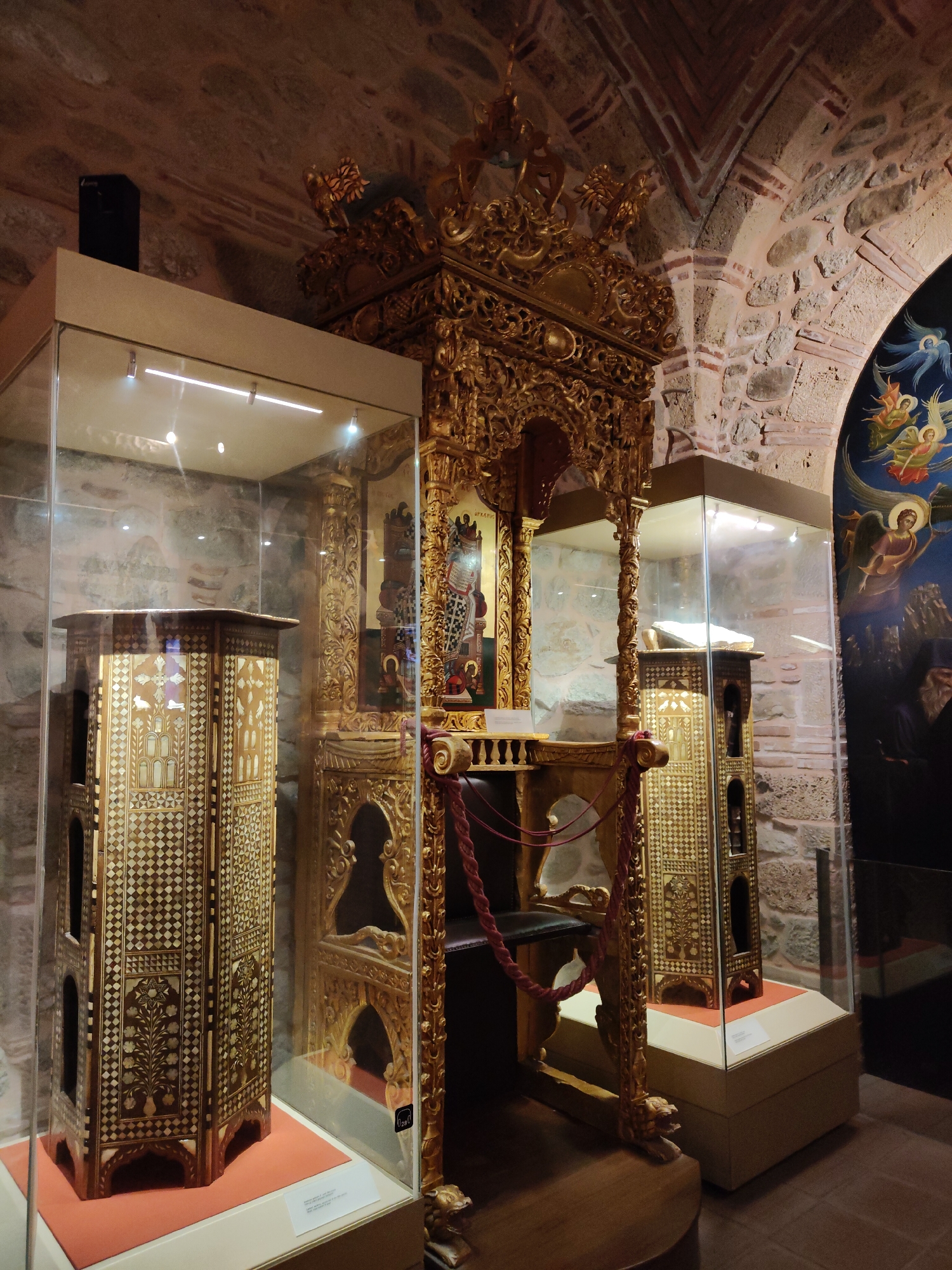 The image depicts the interior of a museum exhibition, likely within the vicinity of Kalambaka, Greece, given the context of monastic treasuries often found in the region. The scene showcases religious artifacts displayed in glass cases and as standalone exhibits, set against a backdrop of ancient stone and brick architecture, characteristic of historical structures in the area.
The primary focus is a large, ornate golden structure, possibly an iconostasis or a bishop's throne, richly decorated with intricate carvings and religious imagery. To the left of this central piece, a tall, hexagonal display case houses a wooden object featuring intricate geometric and floral patterns inlaid with lighter material. This object appears to be a decorative lectern or a reliquary. To the right, another glass case exhibits a similar wooden object with patterned decoration, topped by a book. Behind these exhibits, the rough texture of stone walls and arched brickwork are visible, suggesting an ancient building. On the far right, a section of a fresco or mural is visible, depicting winged figures, likely angels, in blue and gold tones. The lighting is dim, with spotlights highlighting the artifacts within the display cases. There are no people visible in the image. The overall impression is one of a well-preserved historical exhibition dedicated to religious art and artifacts.