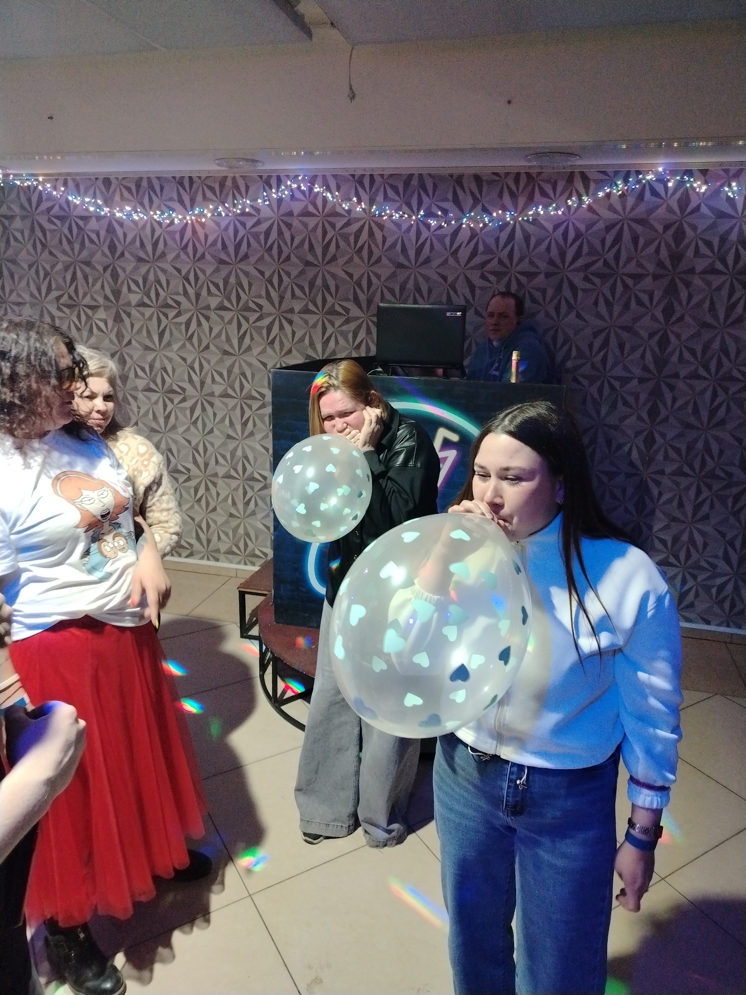 Several people are in a room, likely a nightclub or event space, indicated by the lighting and patterned wallpaper. A woman in the foreground is blowing up a clear balloon filled with small blue hearts. Another woman beside her is holding a similar balloon. To the left, two other women are talking. In the background, behind a DJ booth, a man is seated in front of a laptop. The location is possibly in Chaikovsky, Russia.