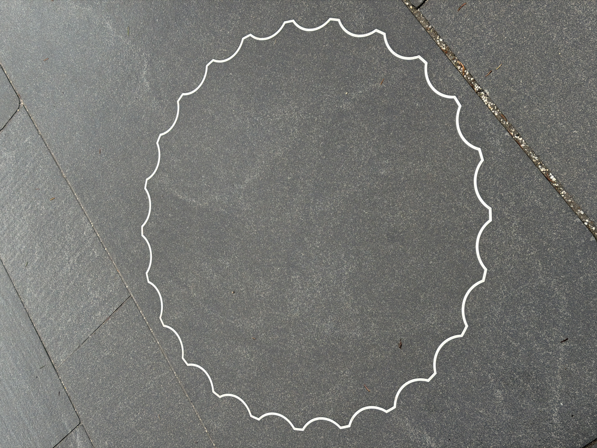 A high-angle shot captures a white, scalloped circular outline drawn on a dark gray, textured tiled surface, likely an outdoor pavement or patio in Moscow, Russia. The circle is centrally positioned within the frame, with its decorative, undulating edge creating a distinct boundary.
The surface consists of large, rectangular dark gray tiles, which exhibit a granular texture and subtle variations in shade. Visible grout lines run diagonally across the image, breaking the tiled pattern, particularly in the upper left corner. A prominent diagonal grout line in the upper right section contains small, light-colored pebbles and debris. Scattered across the tiled surface, both inside and outside the white circle, are small pieces of natural debris, such as tiny twigs or leaf fragments.
There are no people or specific activities depicted. The lighting is bright and even, suggesting a clear or overcast daytime environment, as no strong shadows are cast. The image is a static capture of the drawn shape on the ground. No visible text is present.