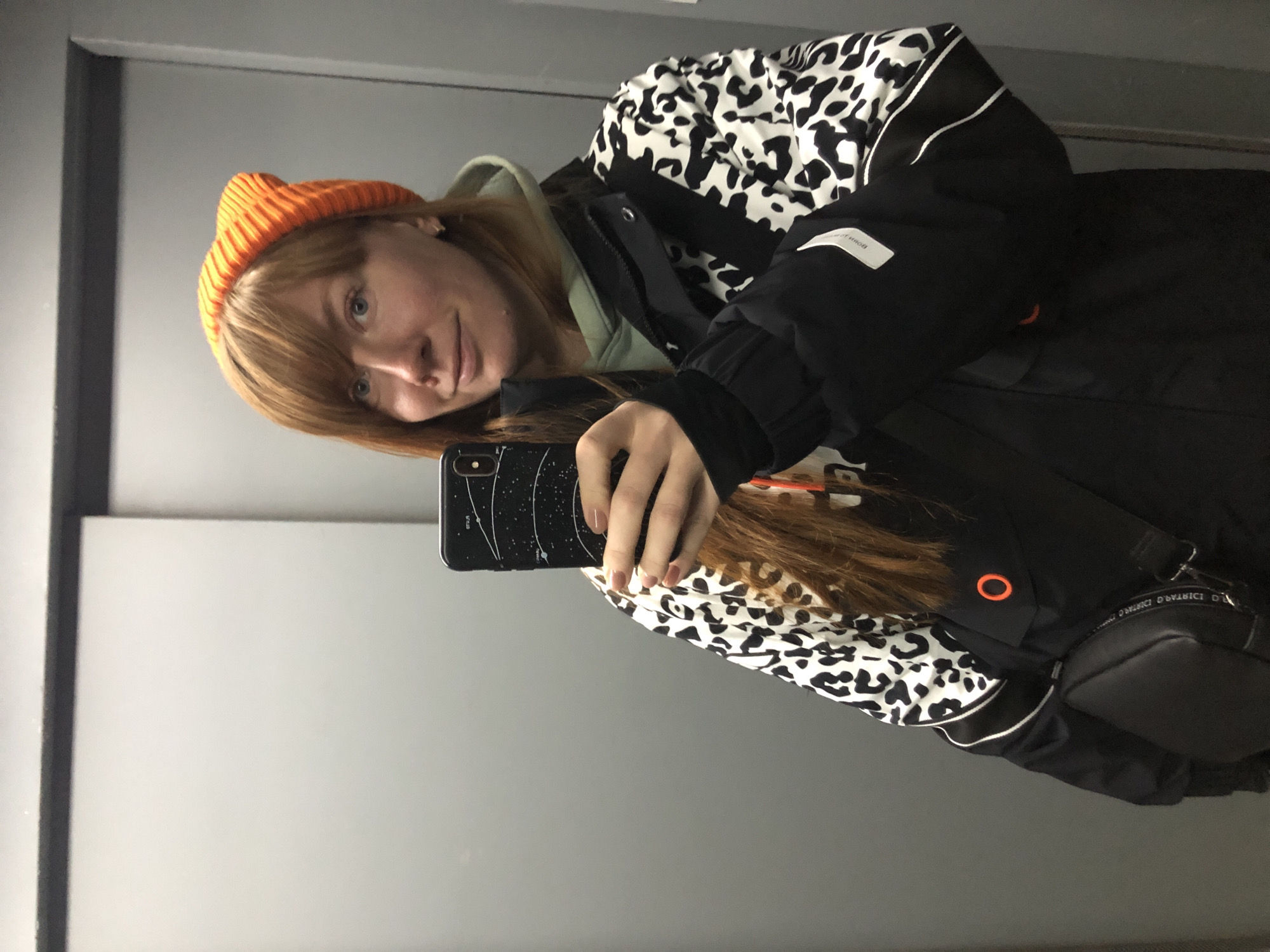 A young woman with reddish-brown hair and an orange beanie is taking a selfie in front of a mirror. She's wearing a black and white patterned jacket and a light green hoodie underneath. She's holding a black smartphone with a constellation design on the back. The background shows a simple grey wall and mirror frame. There is no specific information to identify the exact location within Krasnoyarsk, Russia.