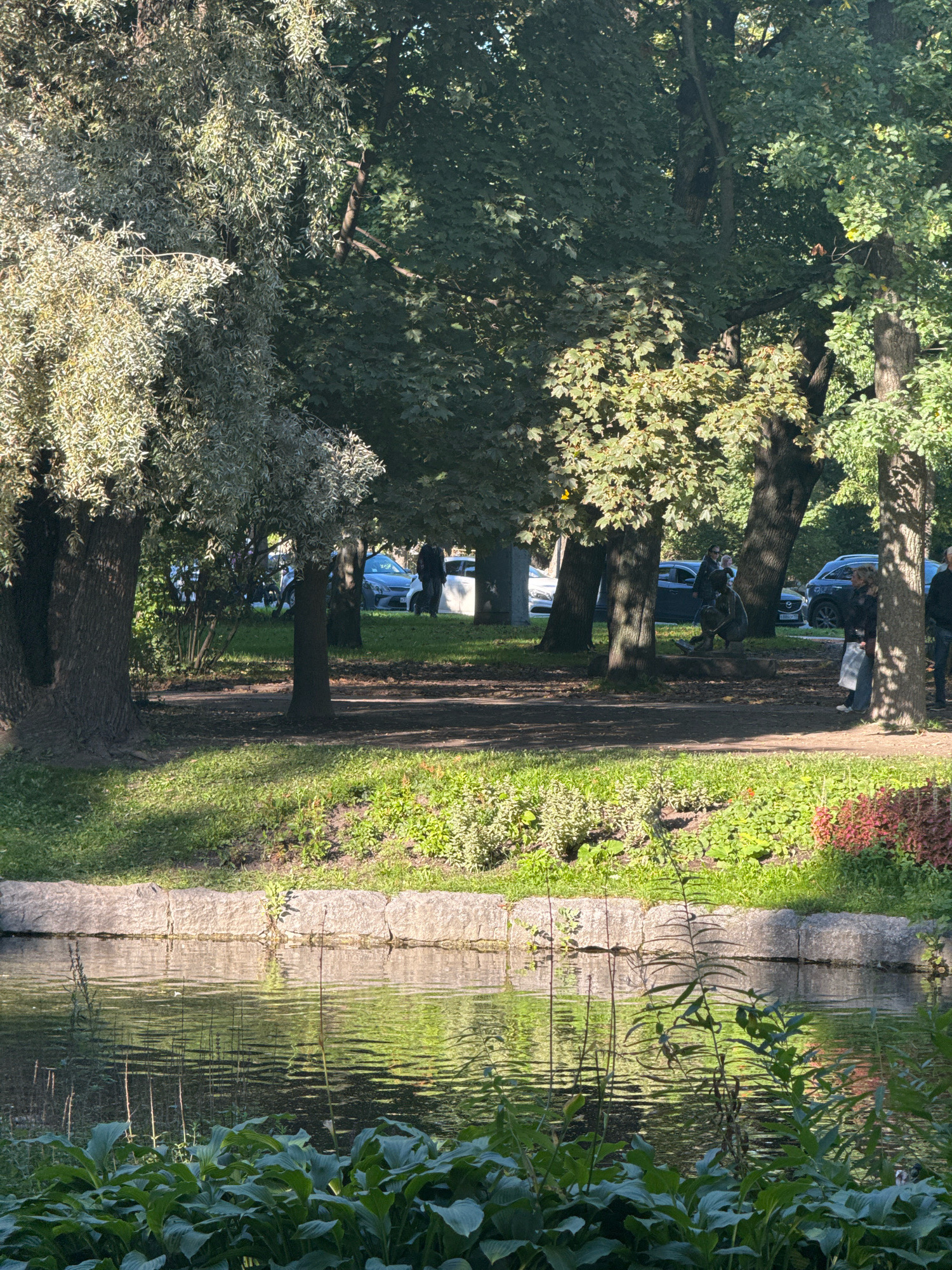 The image depicts a serene park scene in Saint Petersburg, Russia, likely during daytime. In the foreground, a tranquil pond reflects the lush greenery and the overcast sky. The pond is bordered by a stone embankment, with dense foliage of hosta plants in the immediate foreground. Beyond the pond, a pathway meanders through a grassy area shaded by mature trees. Several cars are parked on a street visible in the background, and a few individuals are seen walking or standing, suggesting a public space. The overall atmosphere is peaceful and natural, indicative of a well-maintained urban park.